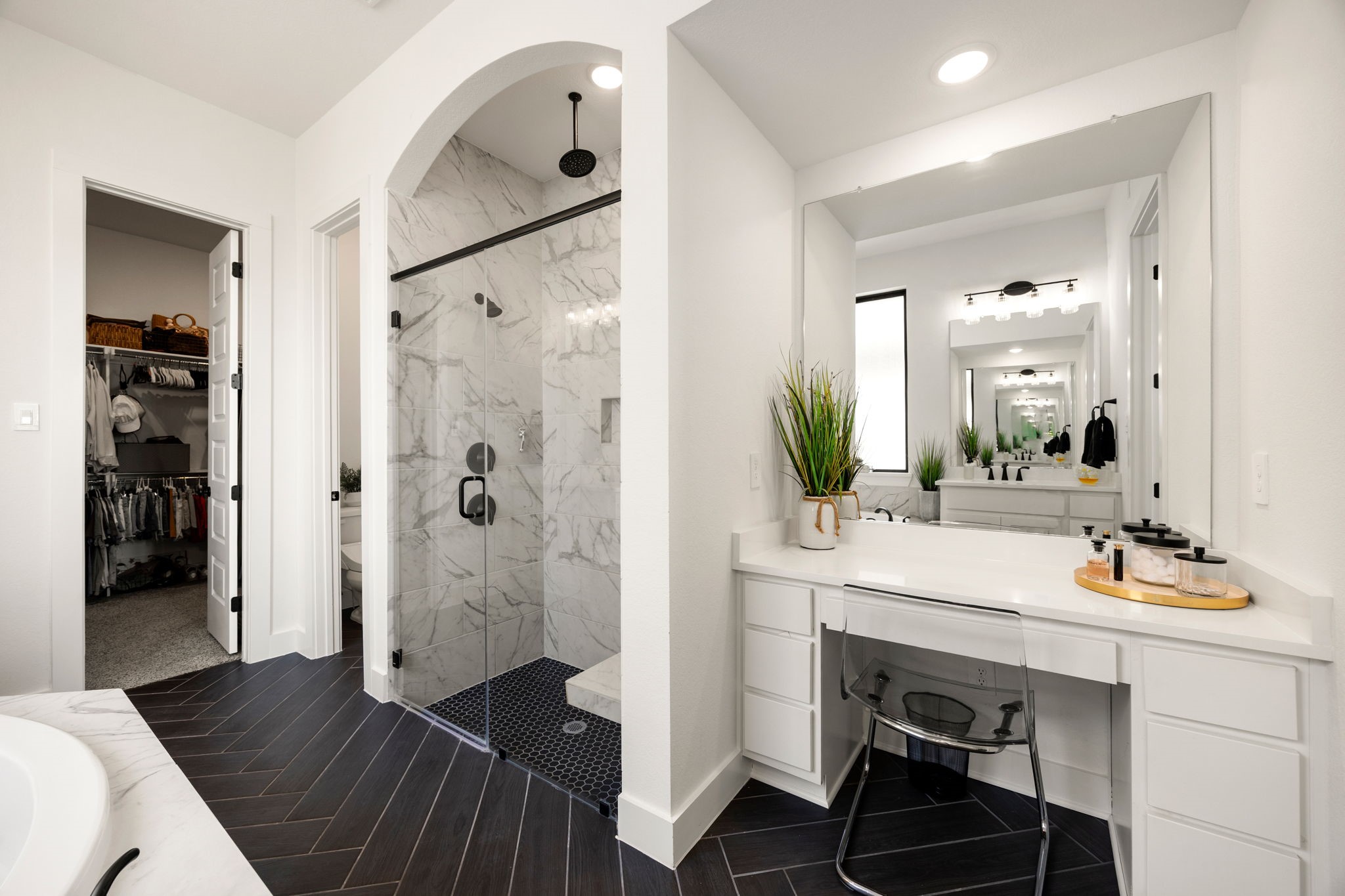 An exquisite custom vanity area. A dedicated makeup vanity with a crisp quartz countertop sits opposite a grand arched walk-in shower featuring seamless glass doors, dual shower heads, and elegant subway tiling, all anchored by rich dark herringbone tile flooring.