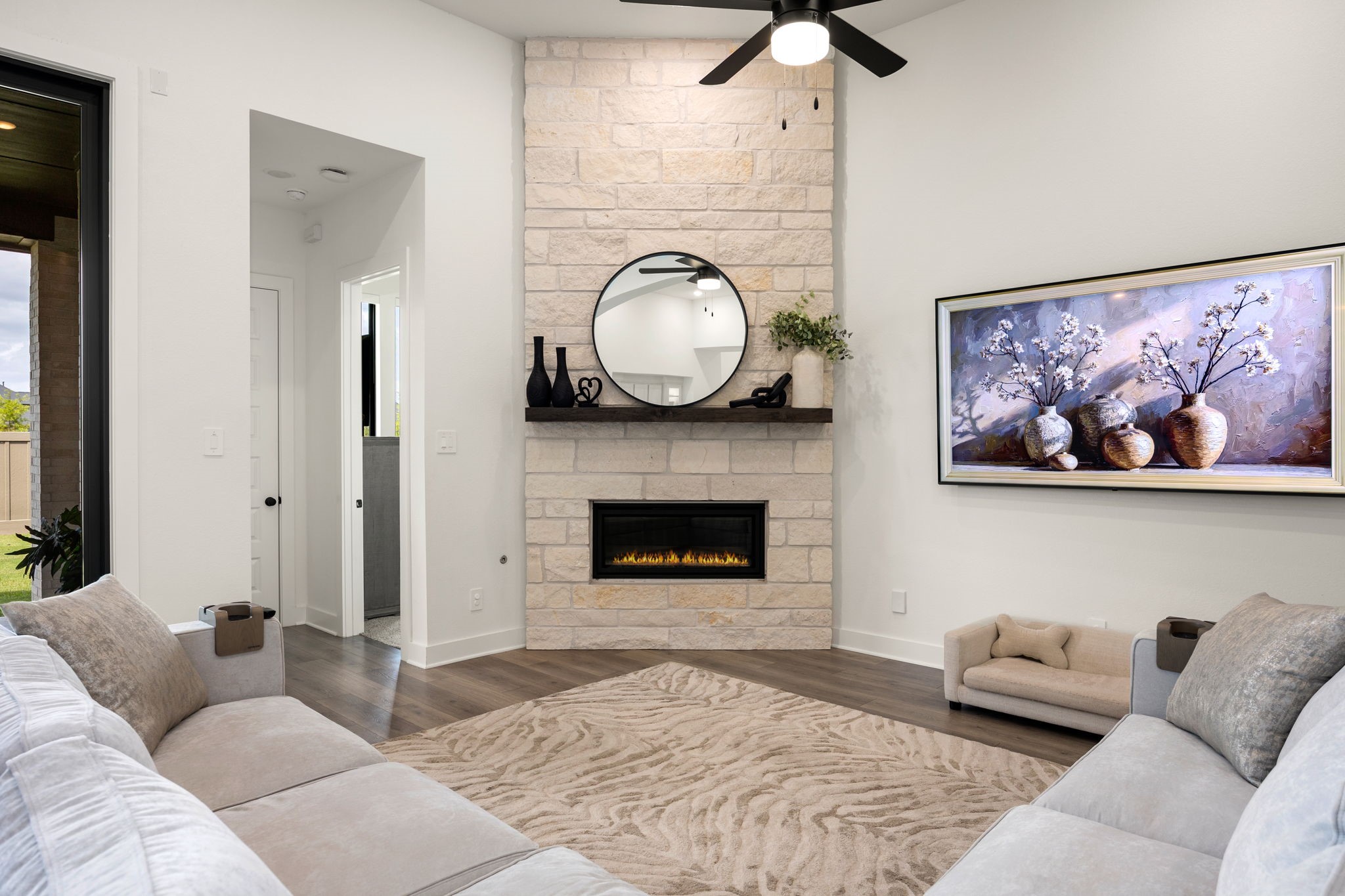 A stunning architectural centerpiece. The corner floor-to-ceiling stone fireplace serves as a gorgeous focal point, beautifully complemented by a dark wood mantel, modern ceiling fan, recessed lighting, and premium wide-plank flooring.