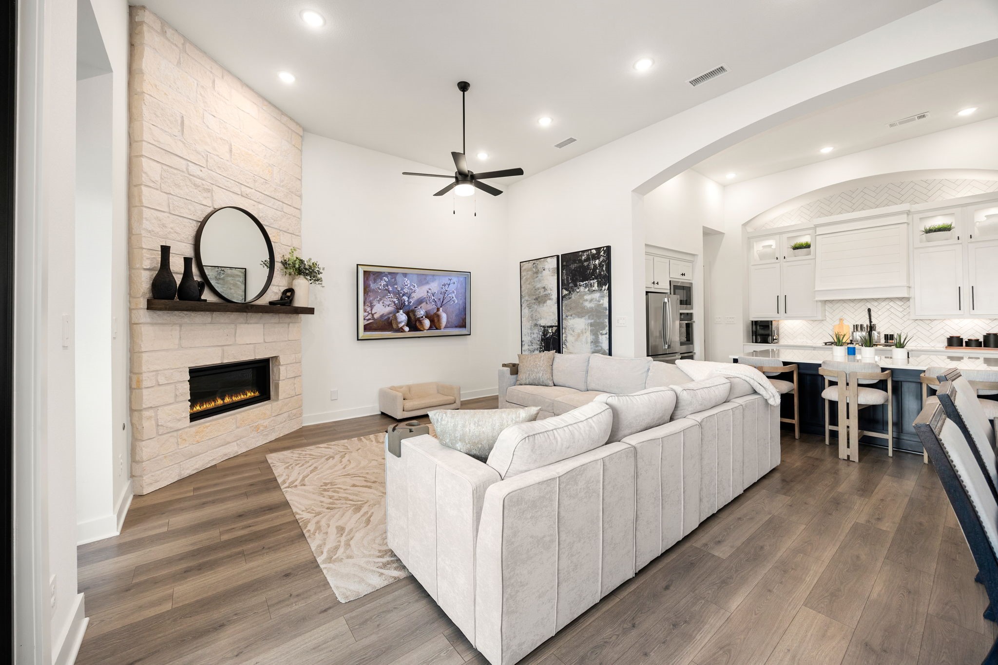 The ultimate open-concept design. High-end wide-plank flooring connects the bright living room to the adjacent kitchen, highlighted by a modern linear stone fireplace, recessed ceiling lights, and an arched architectural walkthrough.