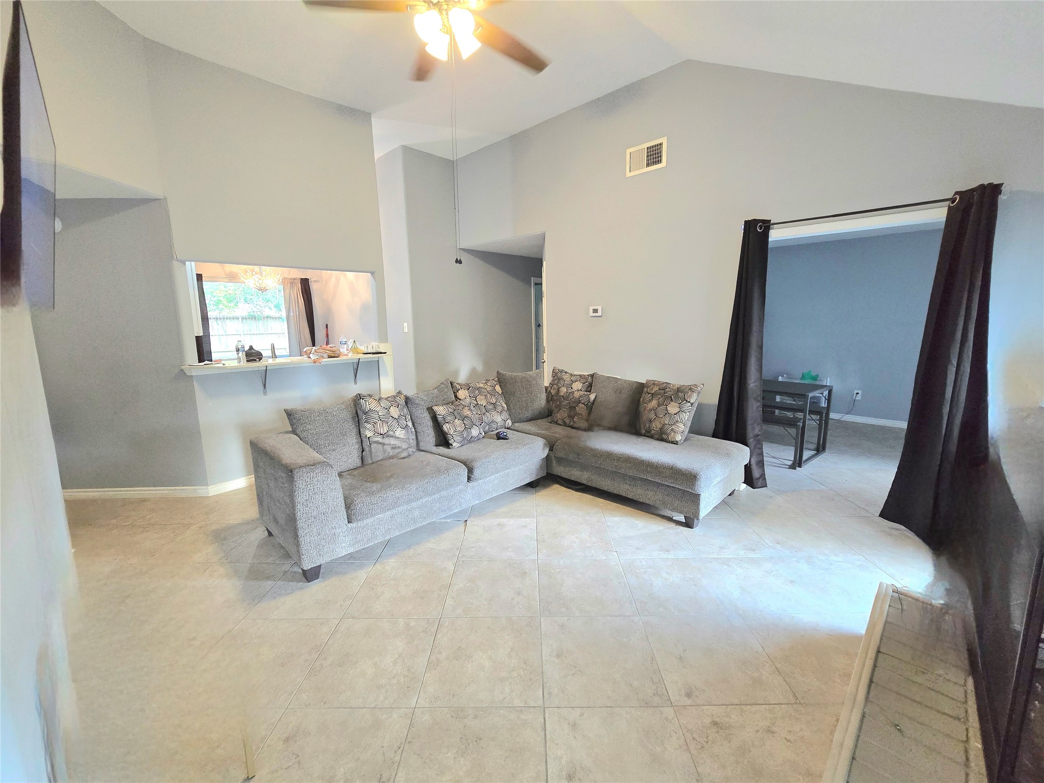 Spacious living area with high ceilings and neutral tones features a cozy sectional sofa, tile flooring, and a convenient pass-through to the kitchen.