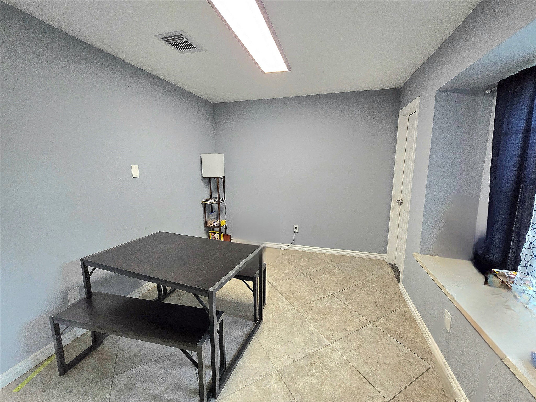 Bright and versatile room featuring neutral tile flooring, soft gray walls, and ample natural light from a large window, perfect for a dining or workspace.