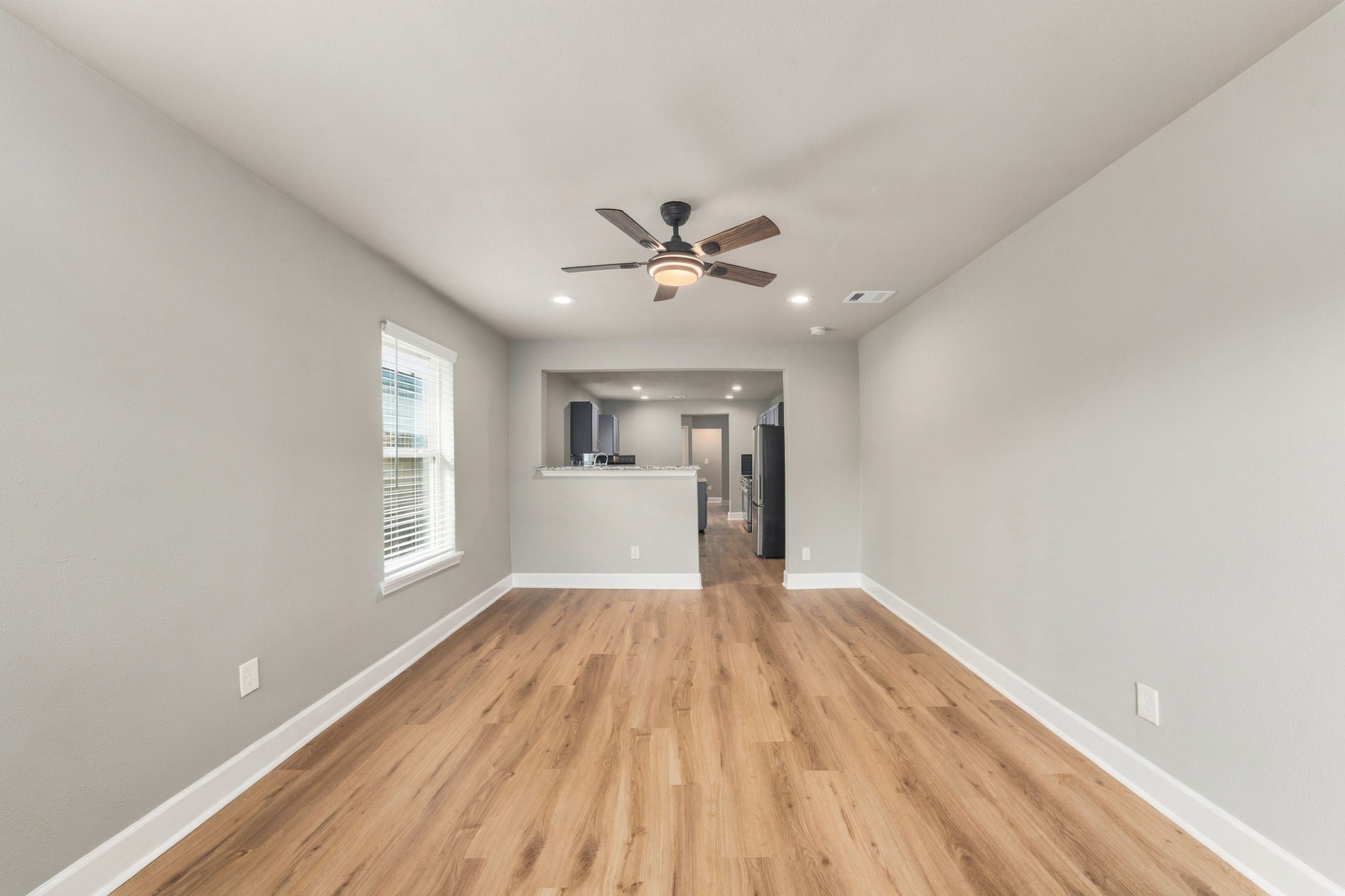 Spacious living area with elegant wood flooring, modern ceiling fan, and ample natural light, seamlessly connecting to the kitchen for an open-concept feel.