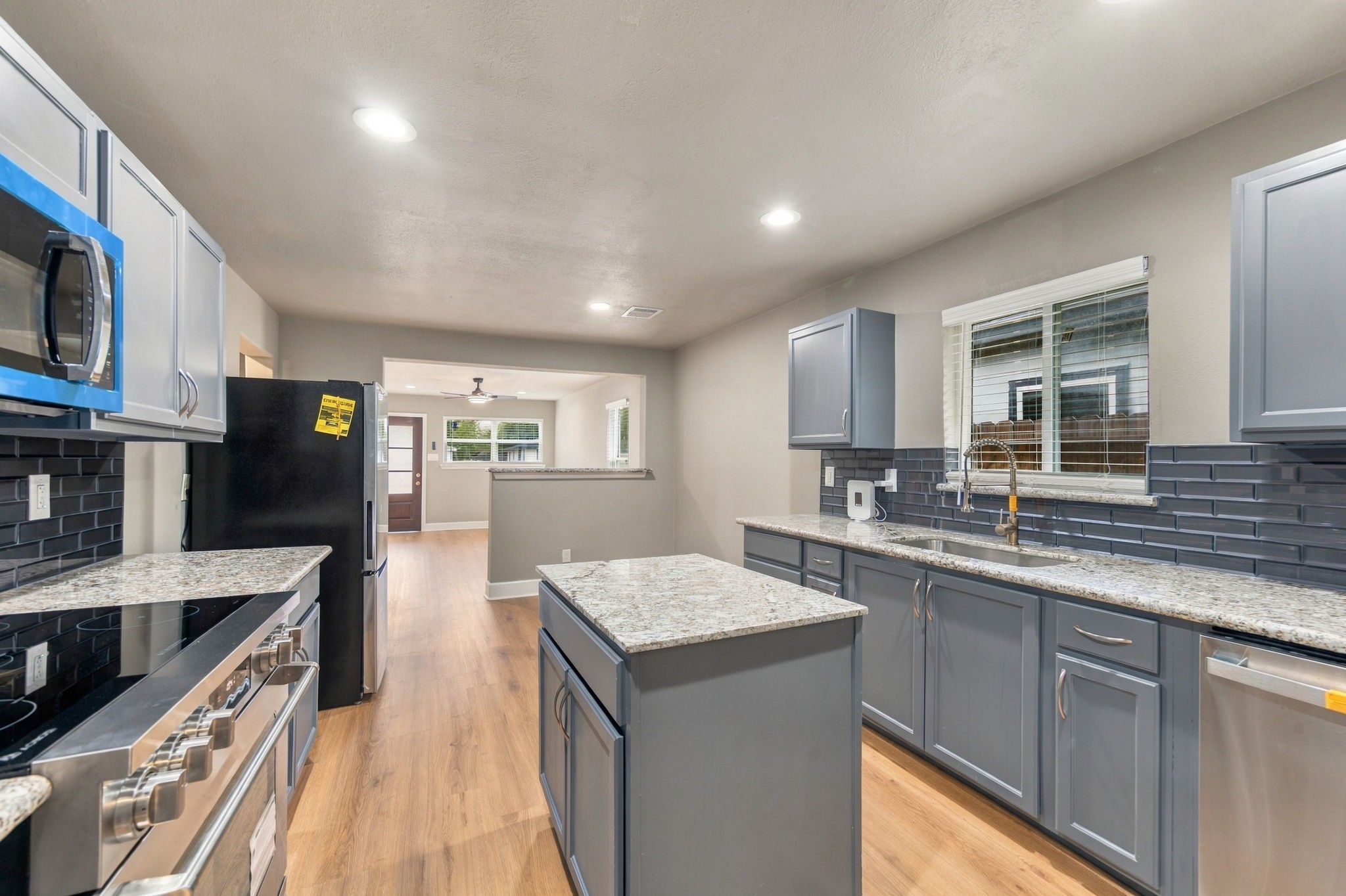 Modern kitchen with sleek gray cabinets, granite countertops, and stainless steel appliances, opening to a bright living area with wood flooring.