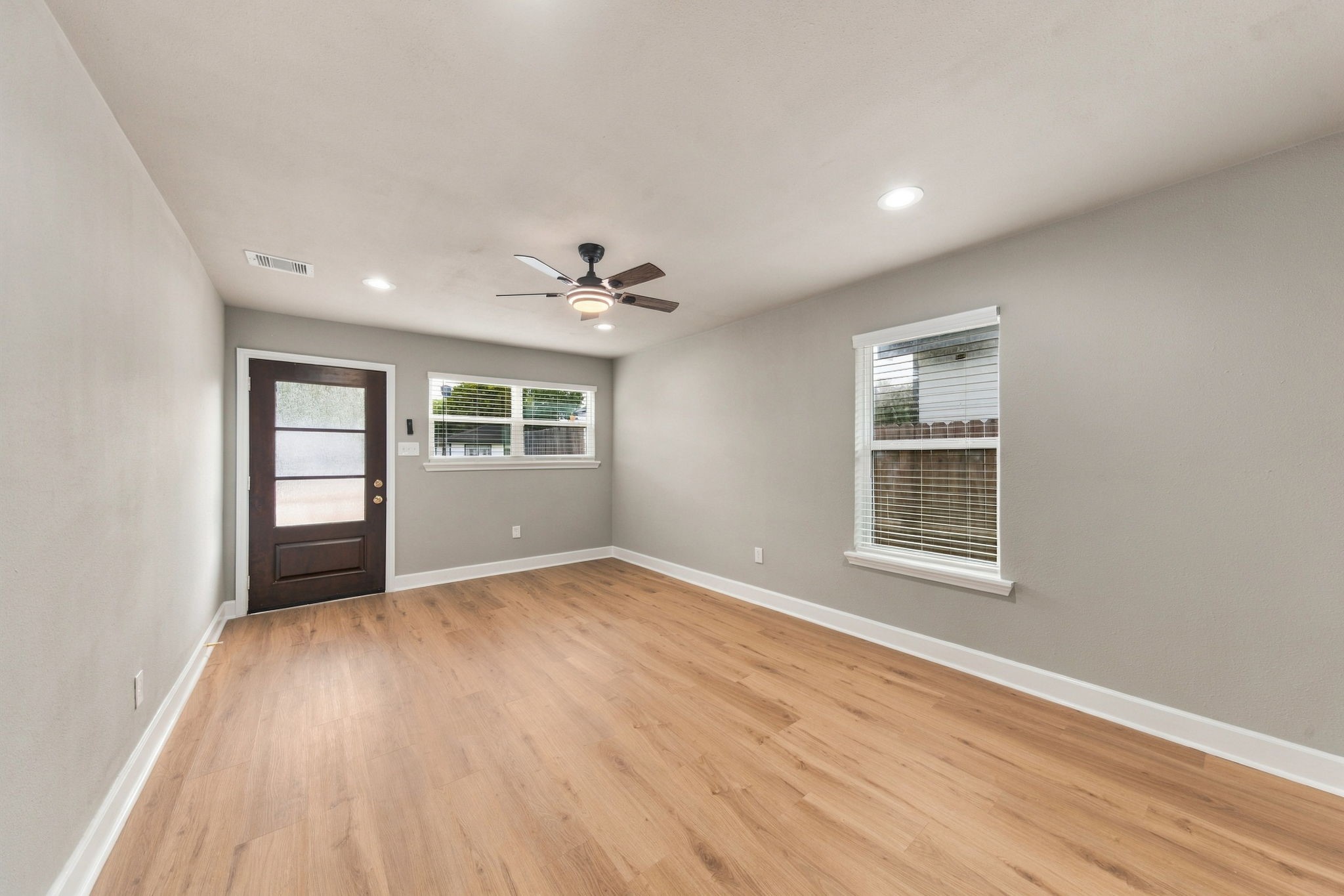 Bright and spacious room featuring sleek hardwood floors, a modern ceiling fan, and ample natural light from two large windows.