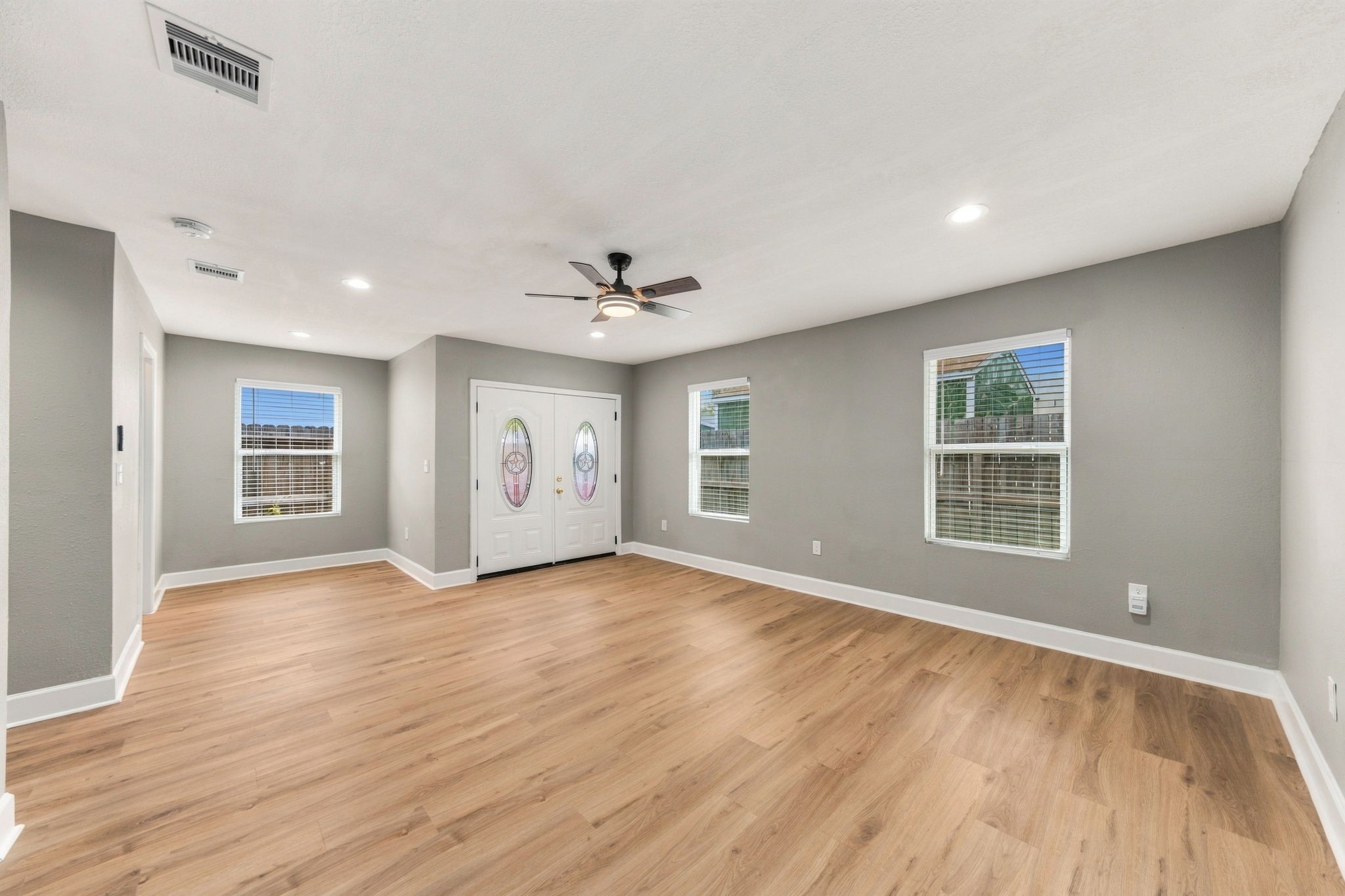 Spacious living area with elegant wood flooring, neutral walls, and abundant natural light from multiple windows, featuring a ceiling fan and stylish double-door entry.