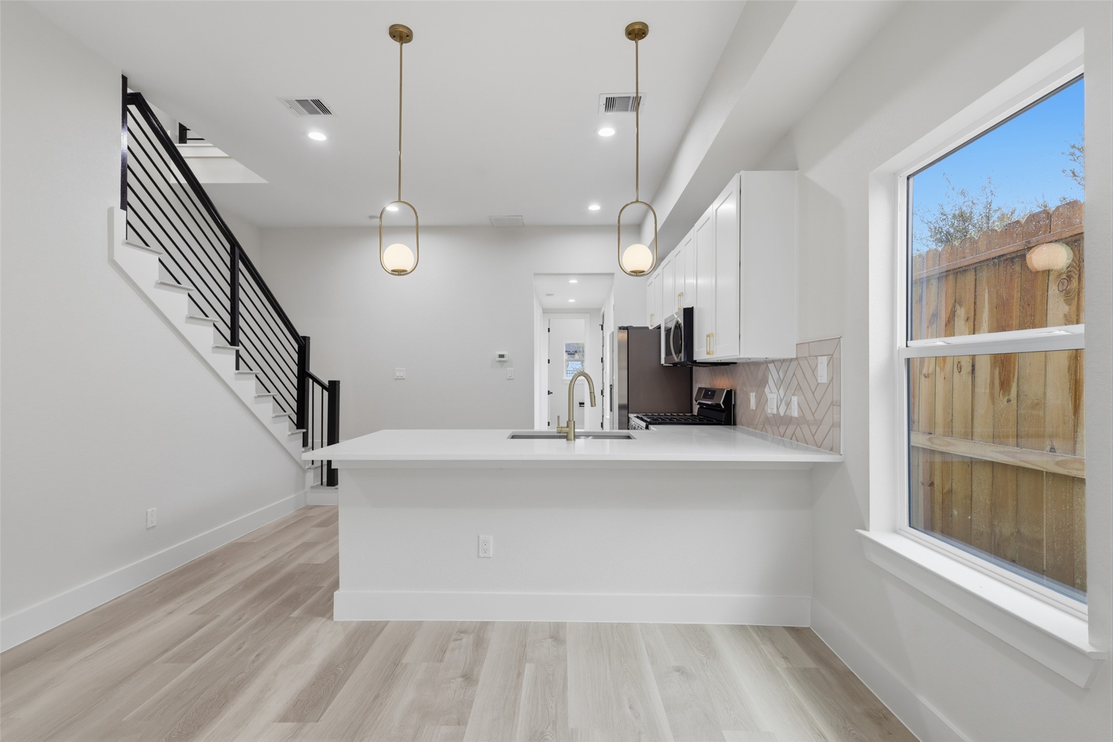 Modern kitchen with sleek white cabinetry, stainless steel appliances, and stylish pendant lighting. The open layout flows into a bright space with a wooden staircase and large window, enhancing natural light.