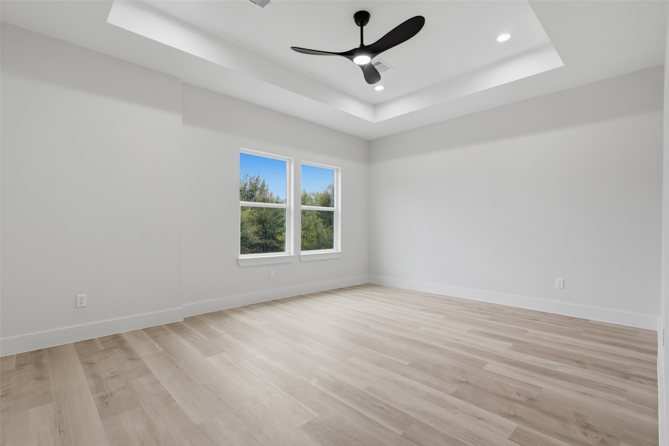 Spacious primary bedroom featuring a tray ceiling with modern fan, abundant natural light from large windows, and sleek LVP flooring.