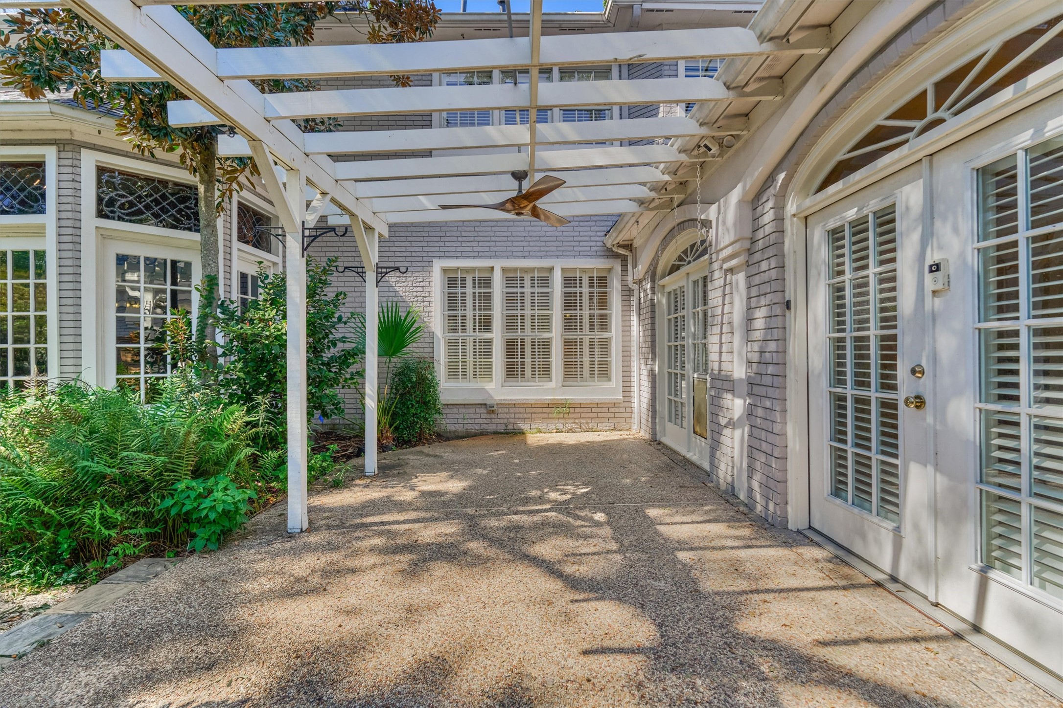 This outdoor patio sits off the sunroom, with a white pergola overhead and a ceiling fan below it. The aggregate floor is framed by brick walls and French doors with plantation shutters and arched transoms, and the librarys leaded glass windows are visible across the courtyard. Mature magnolia, ferns, and a palm soften the edges, and plant hooks on the pergola posts are ready for hanging baskets.