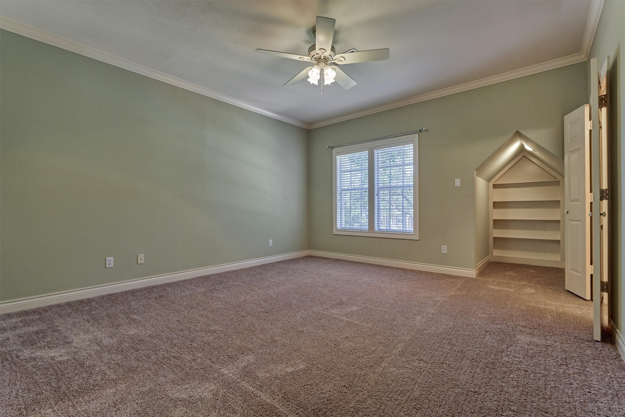 This spacious upstairs secondary bedroom has crown molding, a ceiling fan with lights, and plush carpet. A built-in, house-shaped shelving niche with a peaked, lighted opening adds a charming storage or display feature. A large window brings in natural light, and the large walk-in closet offers plenty of room. The bedroom shares a bath with another secondary bedroom.