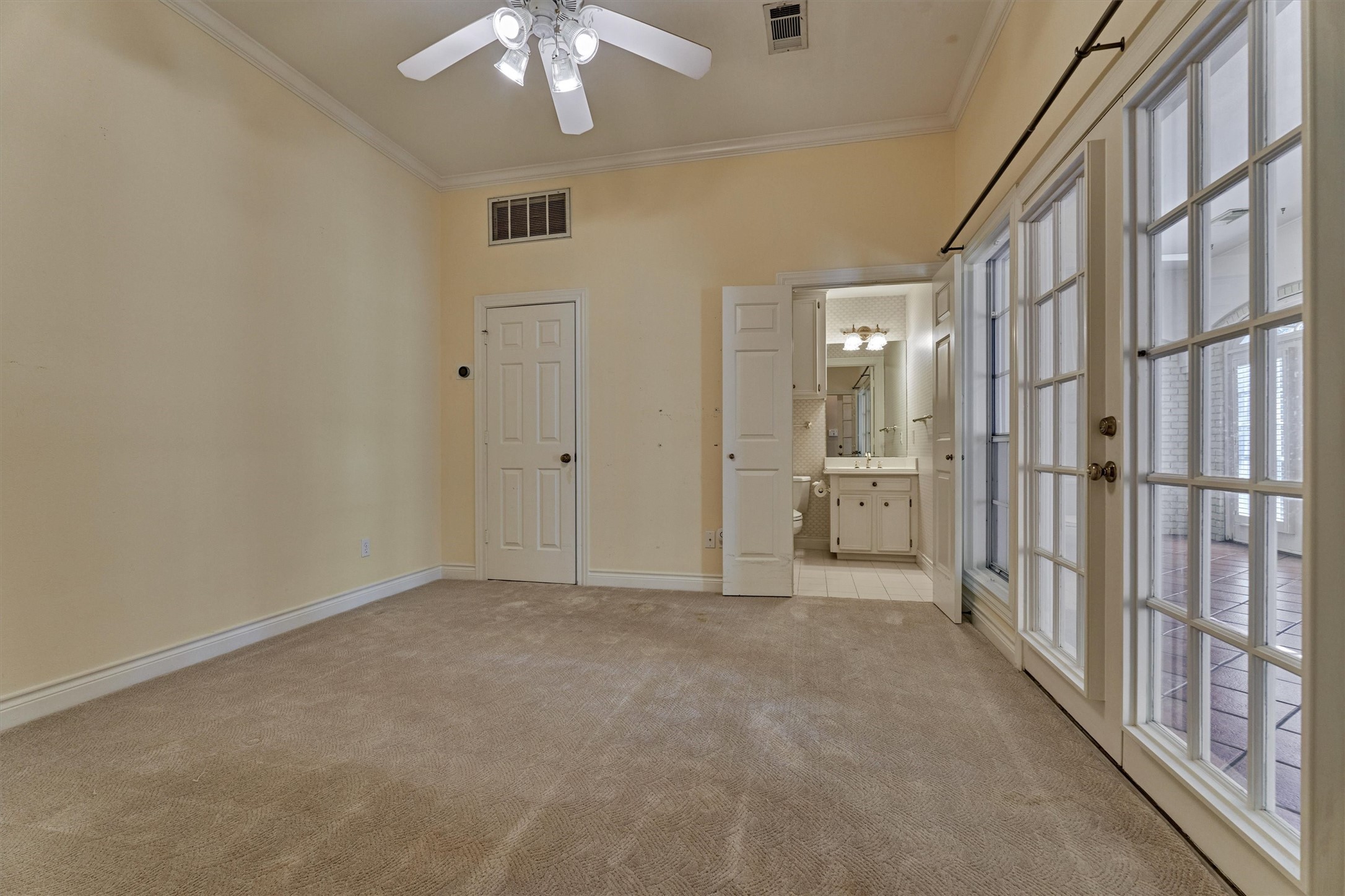 This downstairs bedroom has crown molding, a ceiling fan with lights, and plush carpet. A wall of divided-light French doors opens to the enclosed sunroom, and the ensuite full bath sits just inside the room with a white vanity, cabinet storage, and a tub or shower. A walk in closet adds generous storage. This is a flexible secondary suite that works well for guests or multigenerational living.