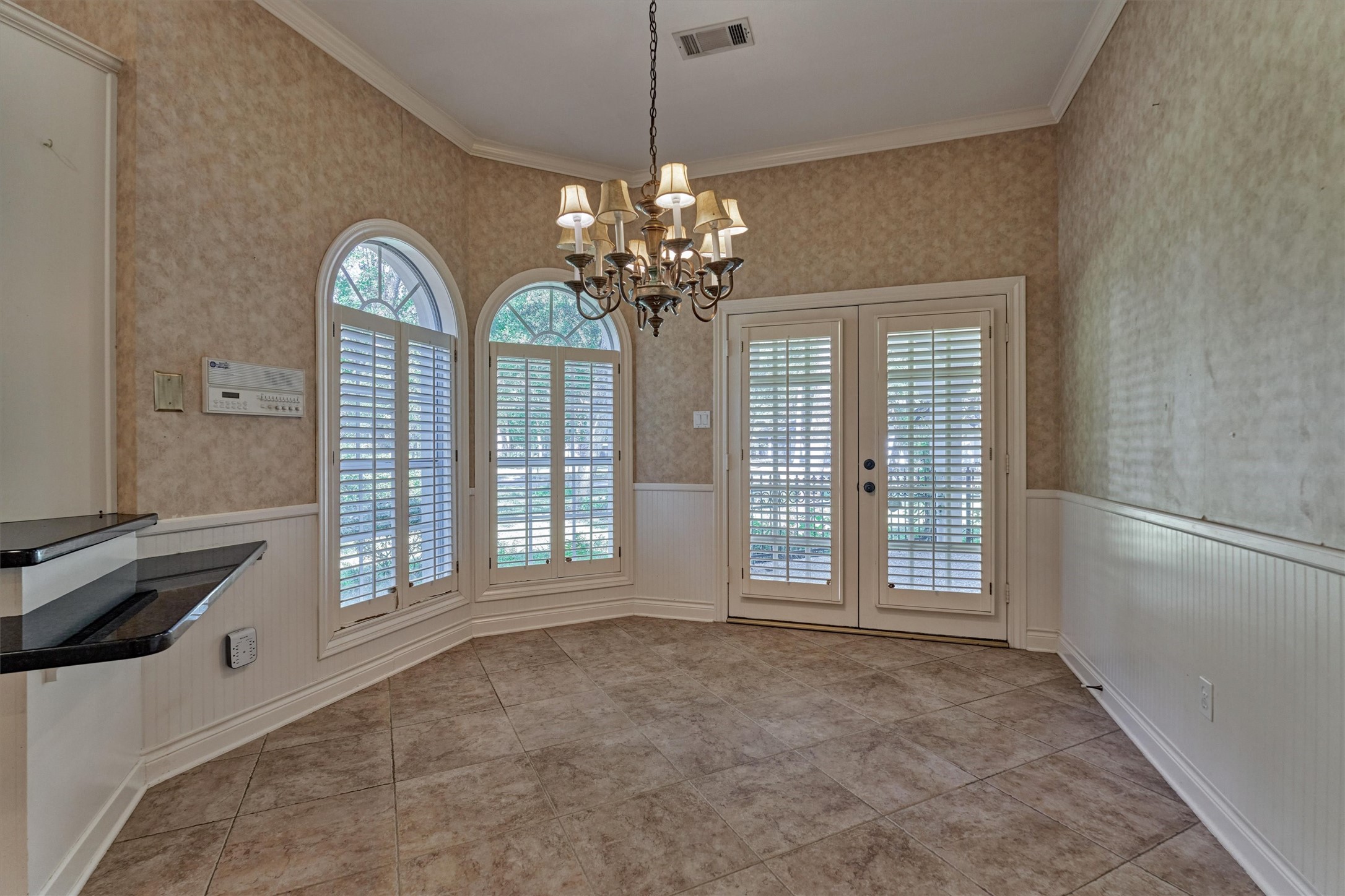 This breakfast area is lit by two arched windows with half-round transoms and plantation shutters, set in a gently angled bay. Beadboard wainscoting with a chair rail and deep crown molding add classic detail, and a chandelier hangs over the tile floors. A granite counter breakfast bar connects the space to the kitchen, and shuttered French doors open to the front covered patio.