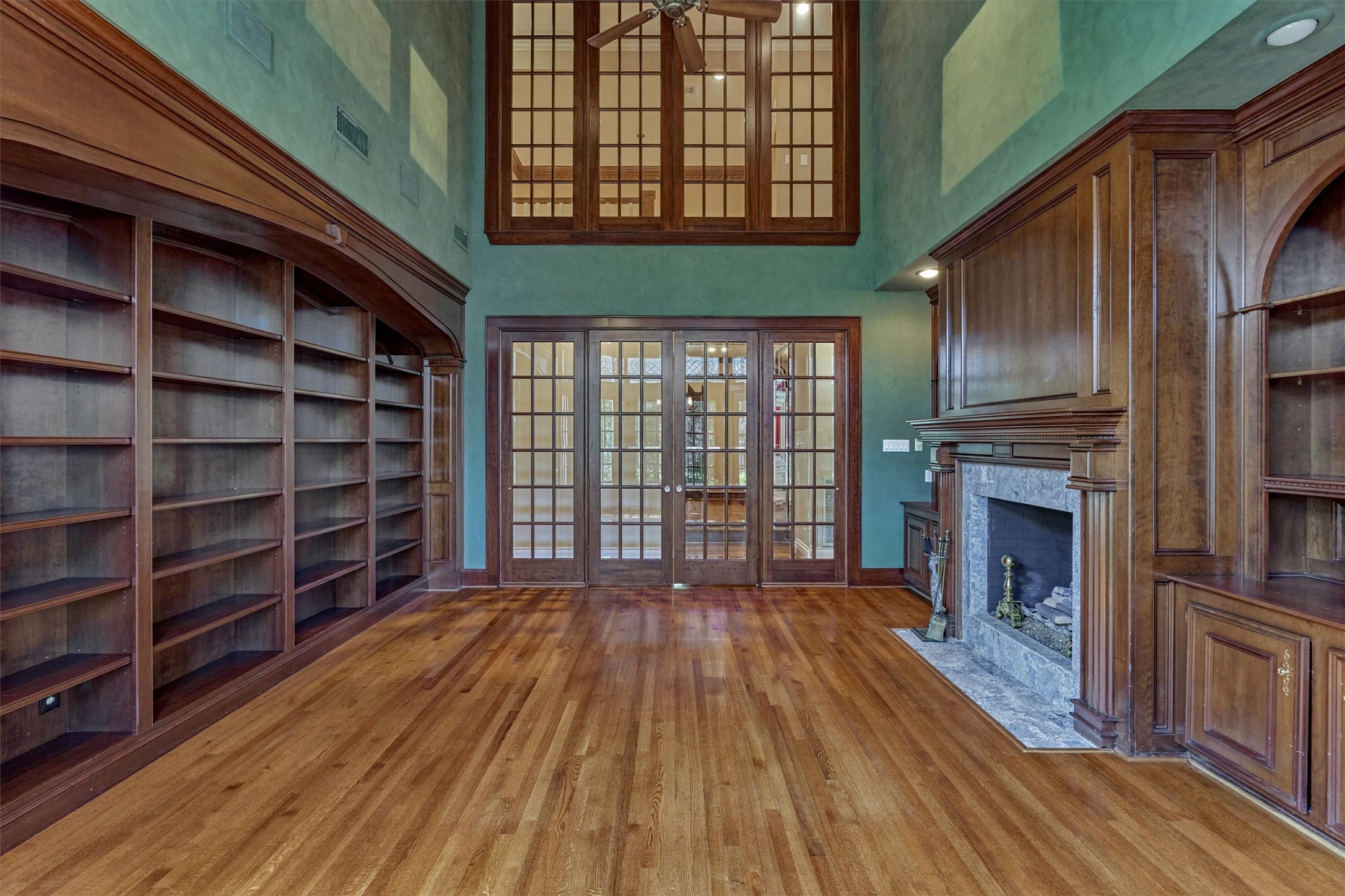 Another view of this two-story library shows its 19-foot ceilings and wall of custom built-in bookshelves beneath an arched cornice. The fireplace wall features raised-panel wood paneling, a carved mantel with fluted pilasters, a granite surround, and an arched display niche. Double French doors with divided lights open to the adjoining rooms, and a high interior window brings light and views between floors.