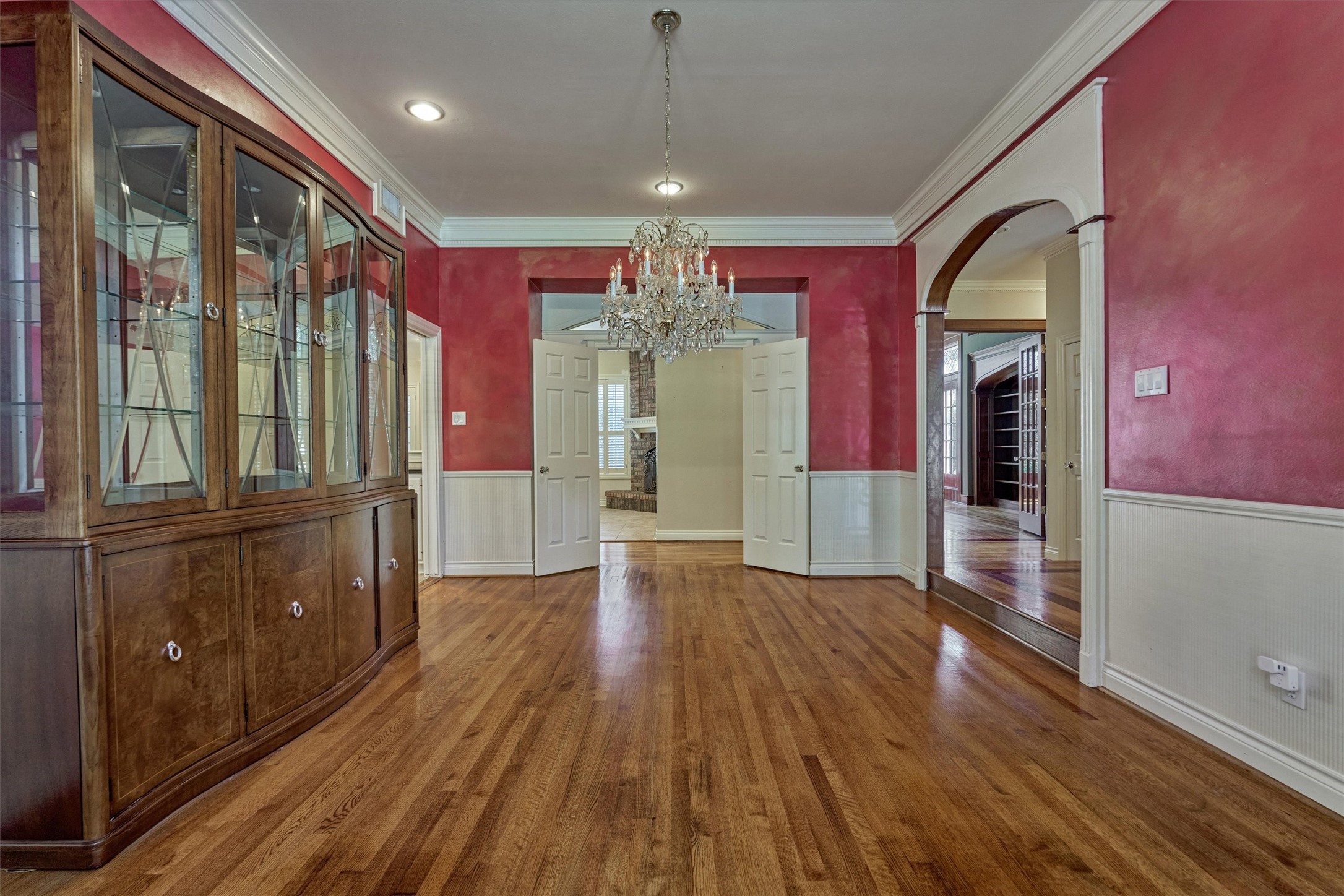 This formal dining room off the foyer has 10-foot ceilings, deep crown molding, and wainscoting with a chair rail. An arched opening trimmed in white leads back toward the foyer, while double doors open to the adjoining family room with its brick fireplace. A crystal chandelier and recessed lighting highlight the hardwood floors, with plenty of space for a large table and a china cabinet.