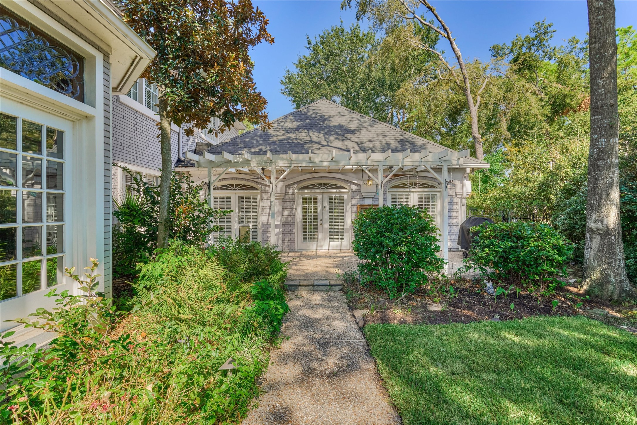 Another angle of the backyard patio shows how easily the house flows outdoors. Three sets of French doors open to a shaded, pergola-covered seating area with room for a grill, dining, and lounging. A winding walkway leads from the lawn to the patio, and tall trees and lush plantings keep it private and peaceful.