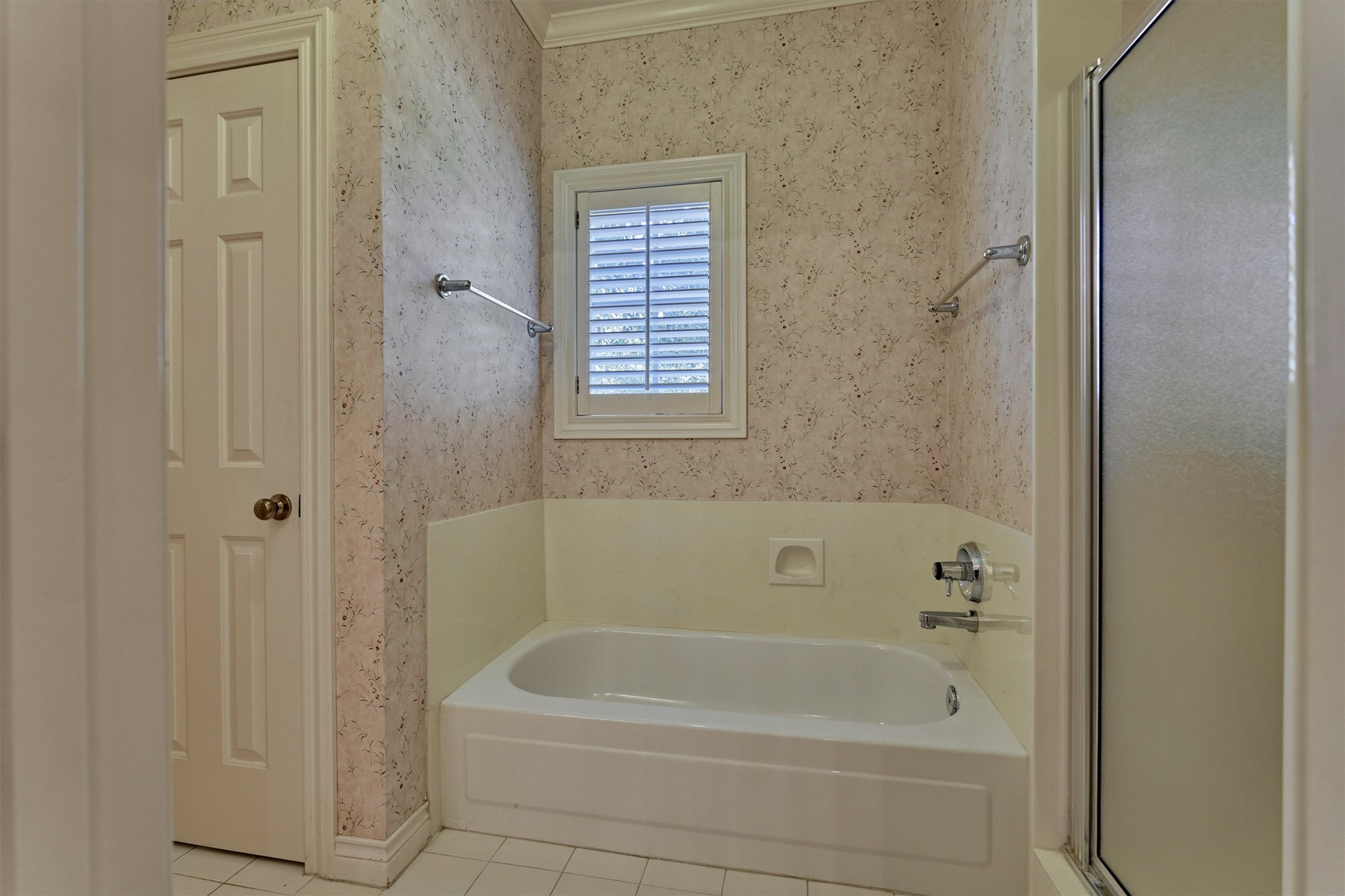 This shared bath has a soaking tub with a tiled-style surround, built-in soap niche, and chrome fixtures, beneath a window with plantation shutters. A separate shower with a frosted glass door sits beside the tub, and crown molding and white tile floors add detail. Doors on either side connect to the private sink and counter areas, so each bedroom has its own space.