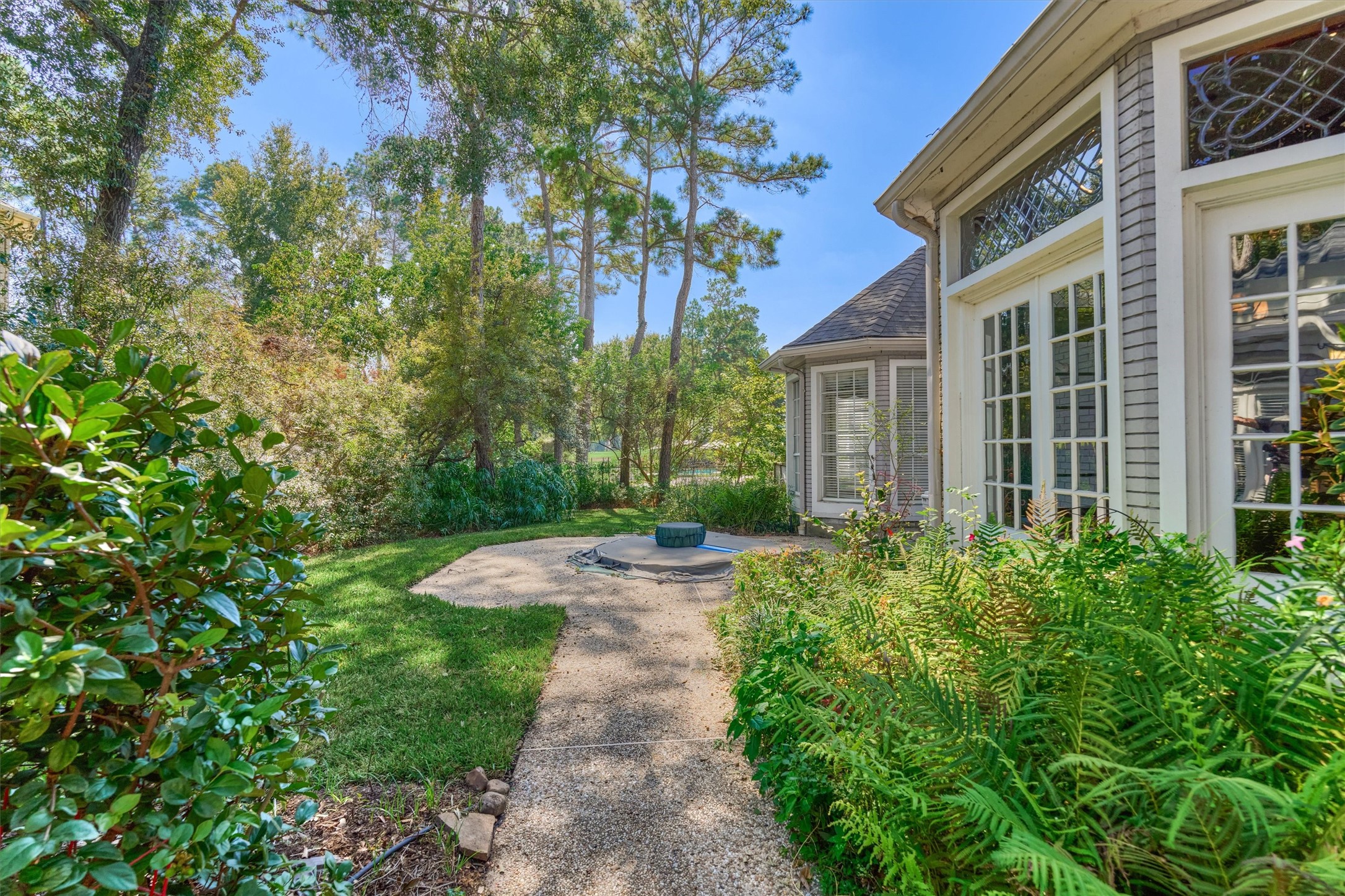 This private backyard has an inground spa on an aggregate patio, with a winding aggregate walkway and a lawn framed by mature pines, oaks, and ferns. The spa is reached from the enclosed sunroom and the primary bedroom through French doors with leaded glass transoms, and a bayed wall of the home adds architectural interest. Through the trees, the golf course community stretches out beyond the back of the property.
