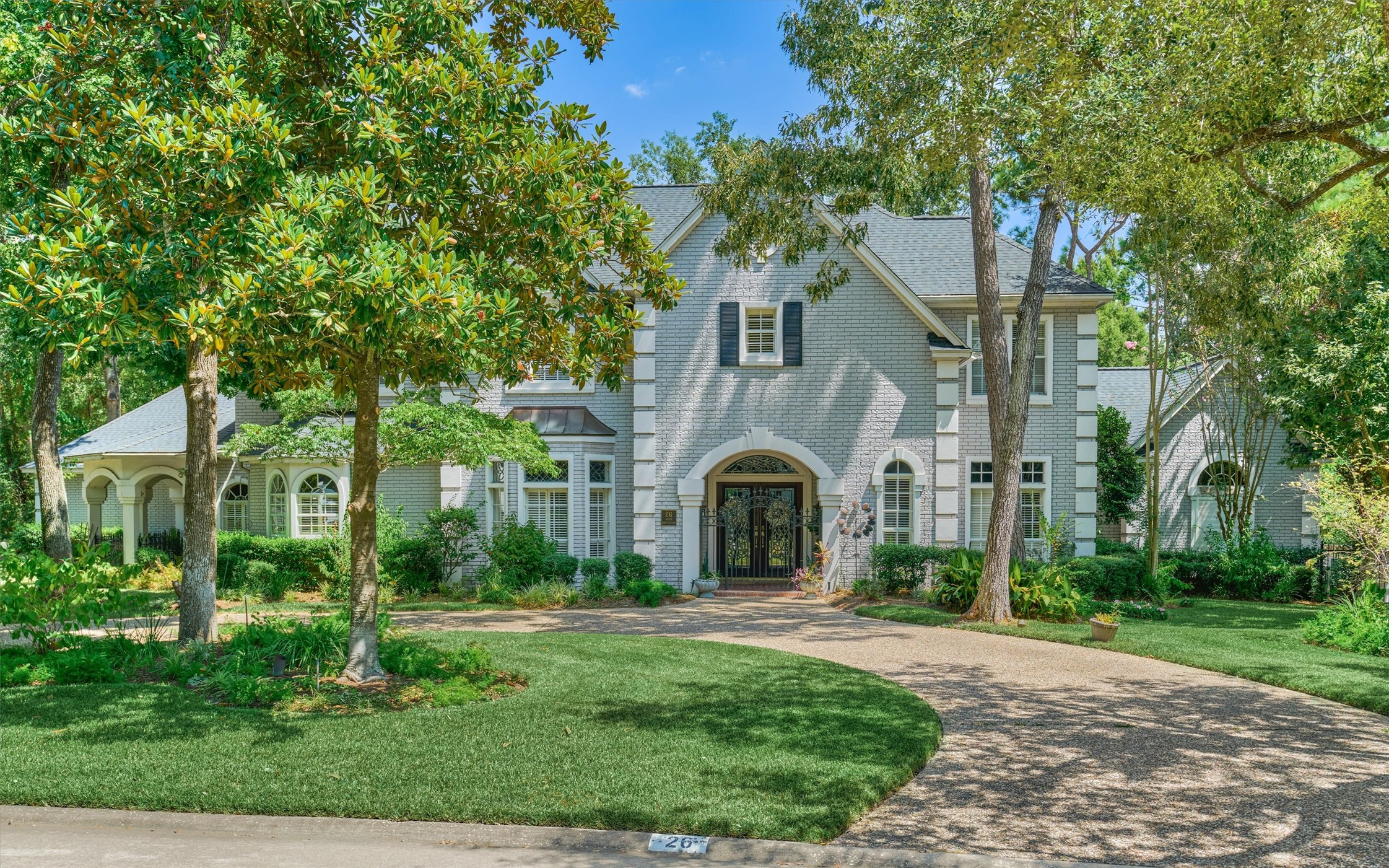 Welcome to 26 S Brokenfern. A circular driveway leads to this elegant brick and stucco home, framed by mature magnolia and oak trees. Arched details, an iron-and-glass entry, and lush landscaping give it timeless curb appeal.
