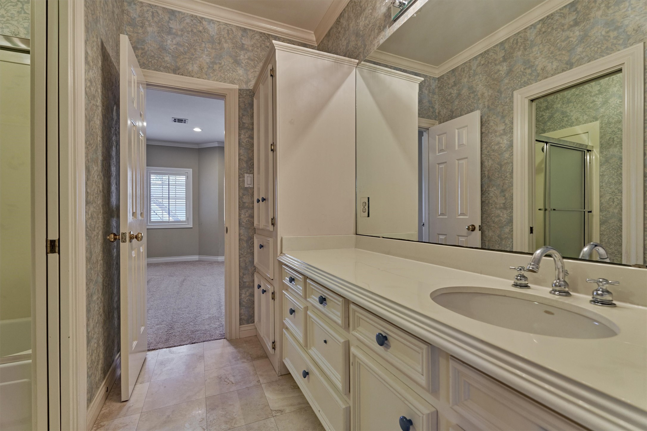 This upstairs bath is a Jack and Jill, set between the third bedroom and the game room. A long vanity with raised-panel cabinetry and drawers sits beneath a wall of mirror, with chrome fixtures and a tall built-in linen cabinet for extra storage. A tub and shower combination with a sliding glass door, crown molding, and large-format tile floors complete the room, with a door at each end that opens to the carpeted bedroom and game room.