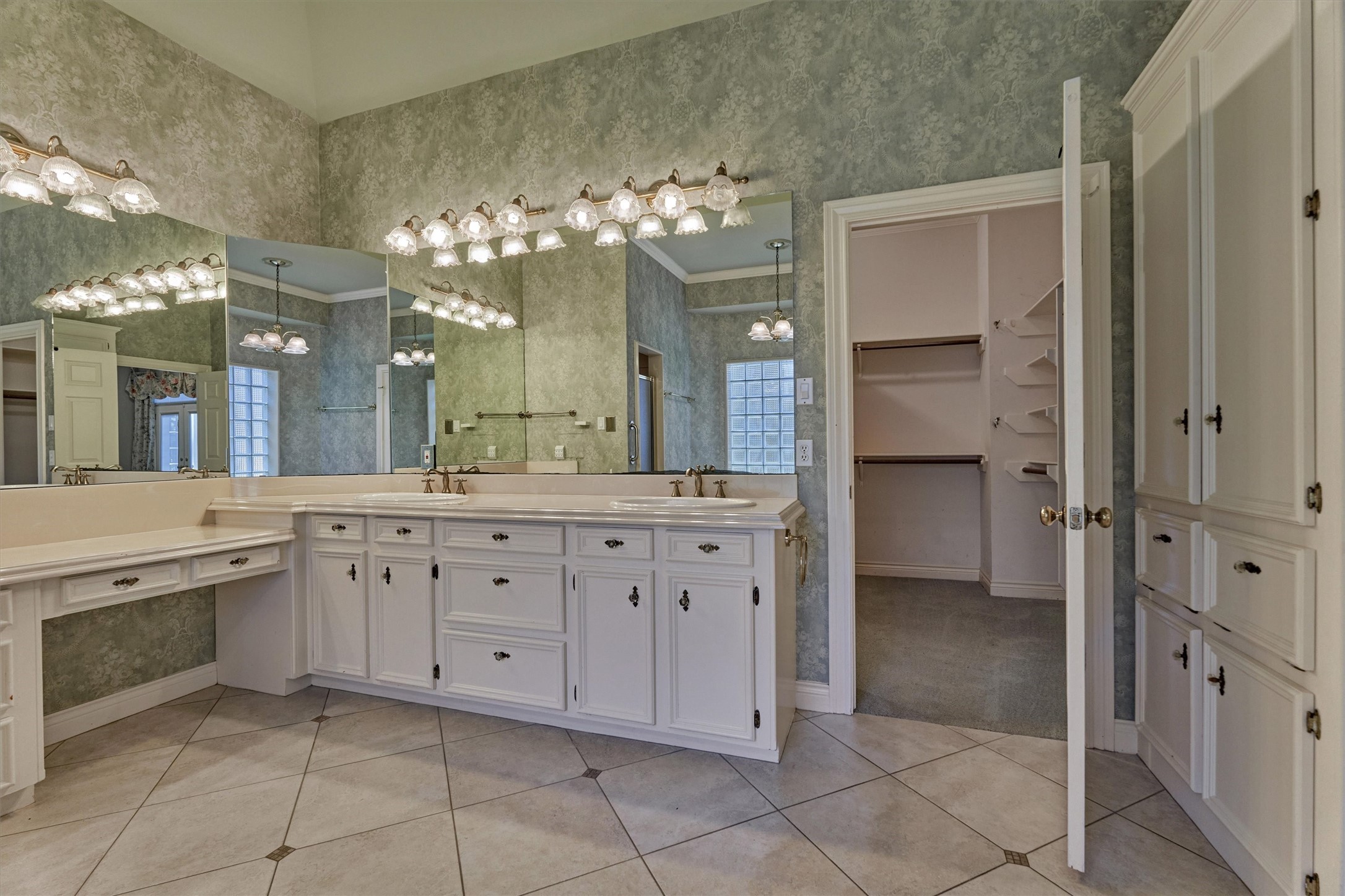 Another view of the primary bath shows a double-sink vanity with raised-panel cabinetry and glass-style knobs, a separate makeup vanity with drawers, and a wall of mirrors with multi-light fixtures. A tall built-in linen cabinet adds storage, and the diagonal tile floor has dark accent inserts. A glass block window brings in natural light. The large hers walk-in closet has built-in hanging rods and angled shelving, and his closet is accessed through hers.