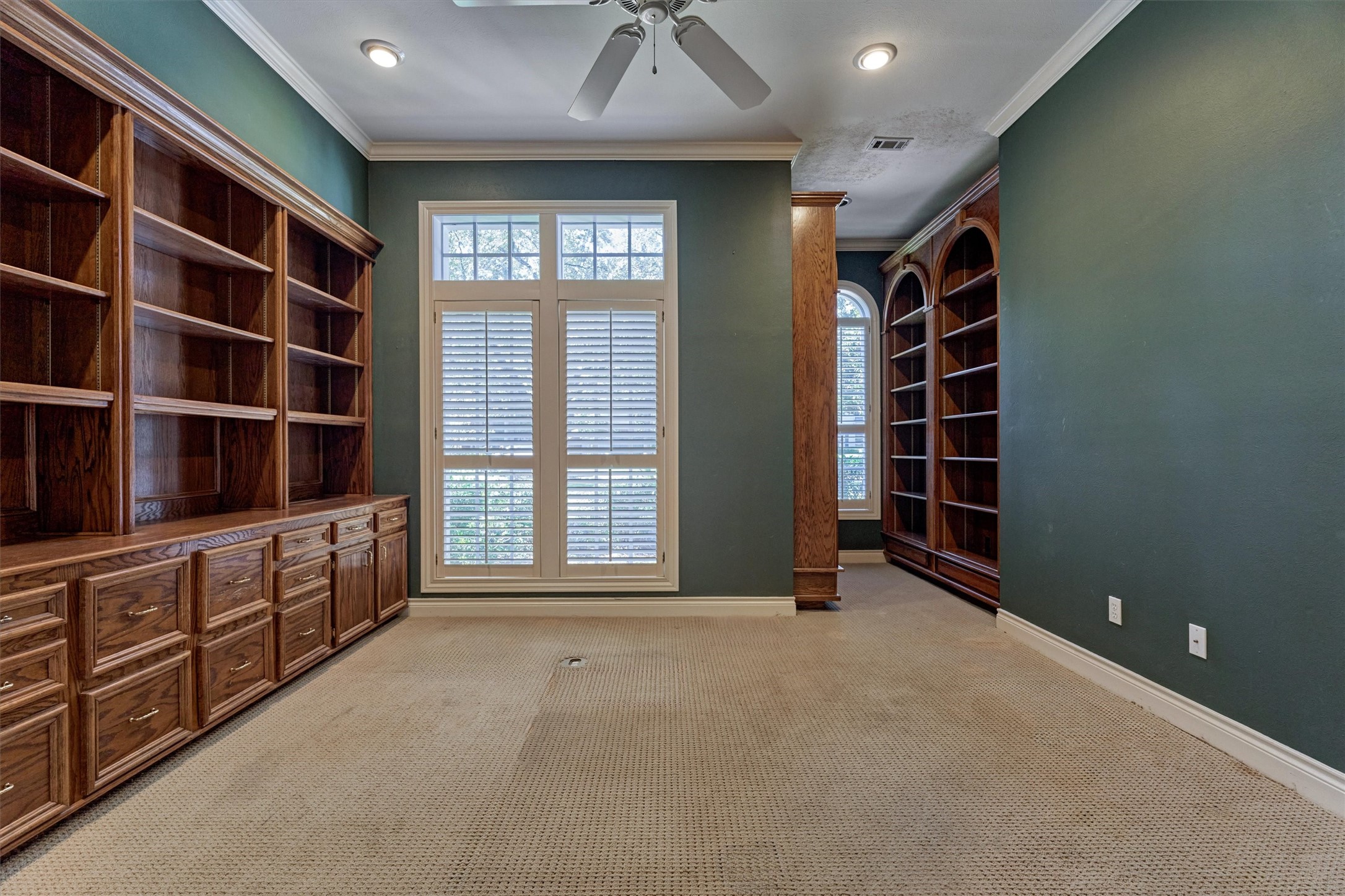 This bedroom is currently set up as a private office, with a full wall of oak built-in shelving and cabinetry that has raised-panel drawers, cabinet doors, and a wide work surface. A tall window with a transom and plantation shutters overlooks the front yard. Crown molding, recessed lighting, and a ceiling fan add detail, and a wood-trimmed column separates a second area with arched, floor-to-ceiling bookshelves and an arched window. A walk-in closet completes the room.