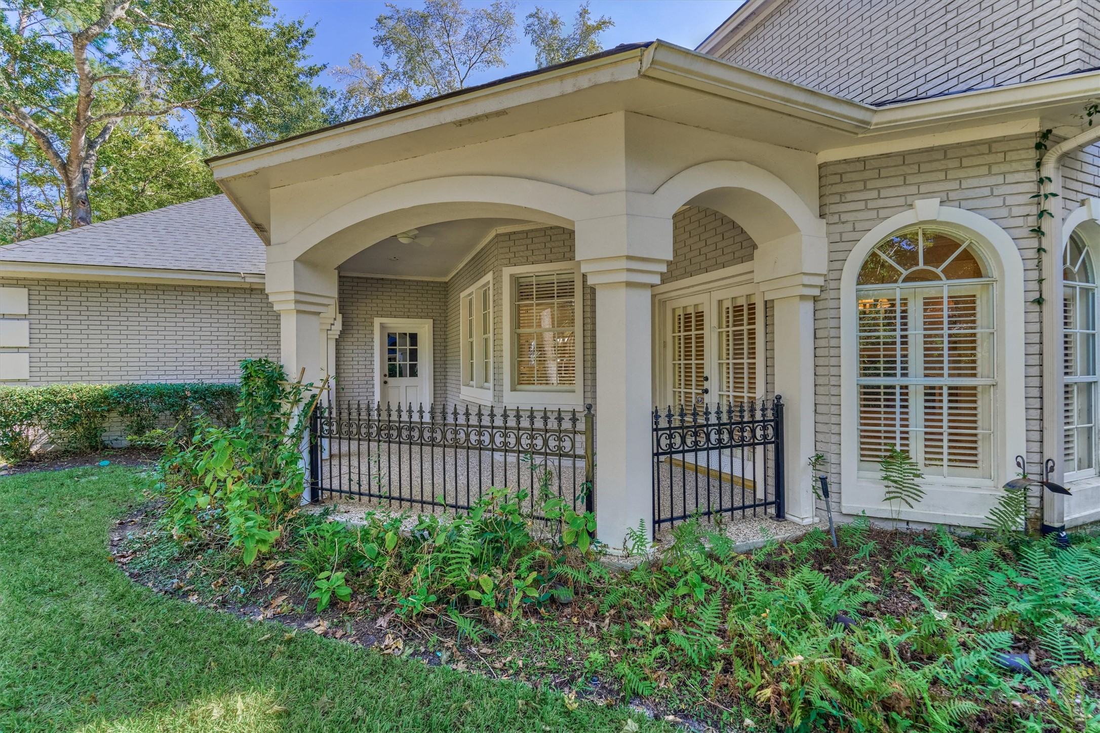 This covered patio sits off the breakfast room and overlooks the front yard, with direct access to both the breakfast room and the garage. Graceful arches rest on stucco columns, with a ceiling fan overhead and a wrought iron railing with scrollwork and fleur-de-lis finials. French doors with plantation shutters open to the breakfast room, and a separate door at the back of the patio leads to the garage. An aggregate floor, brick walls, and an arched window complete the view.
