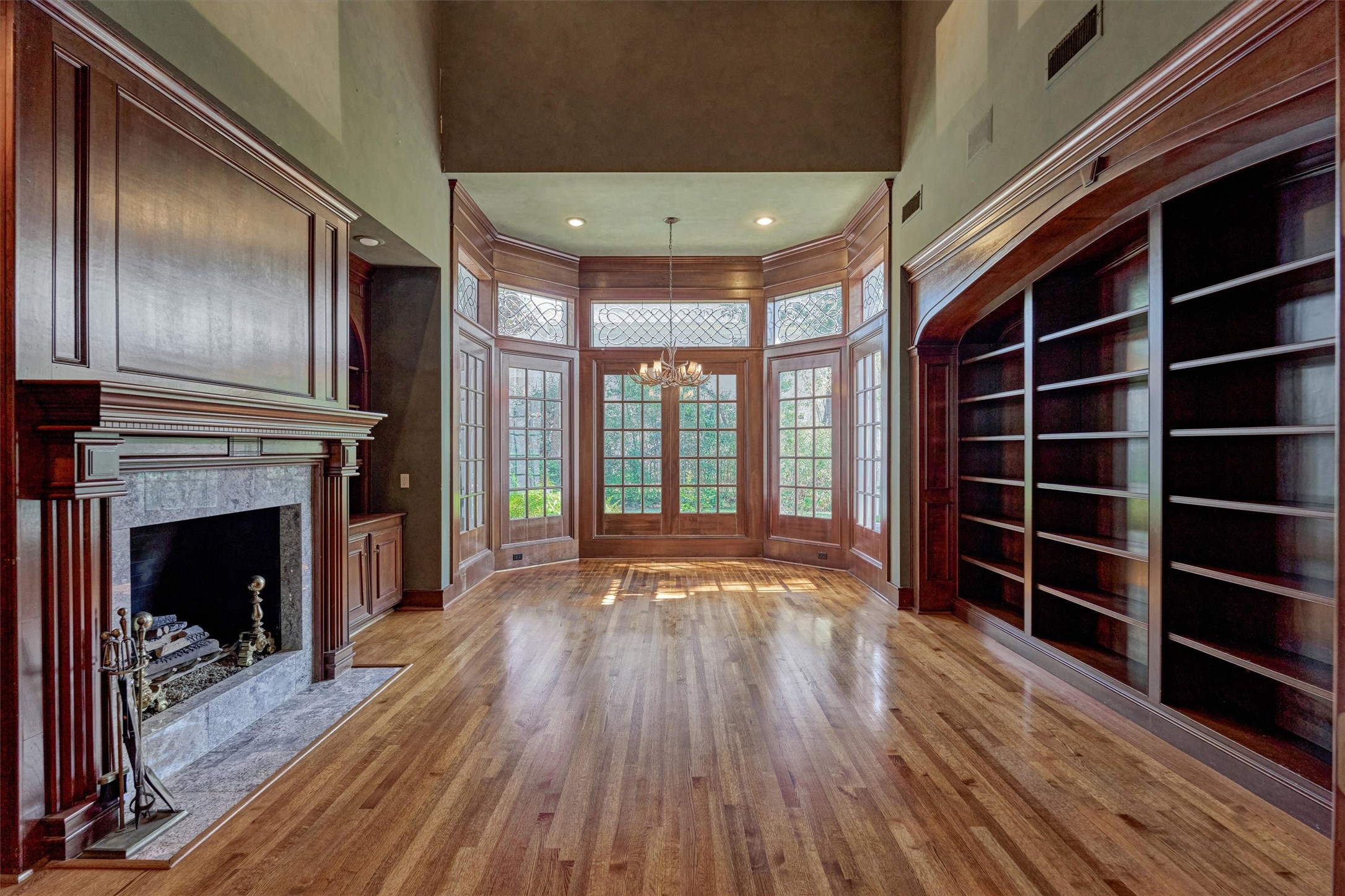 Picture a quiet evening by the fire in this two-story-ceiling library. Floor-to-ceiling cabinetry, a classic wood mantel with dentil molding, and a curved bay of mullioned French doors create an inviting, Ralph Lauren, old-world feel. Recessed lighting and an antler-style chandelier add warmth, and the garden views are a lovely backdrop. Use it as a music room, reading retreat, or executive-style home office.