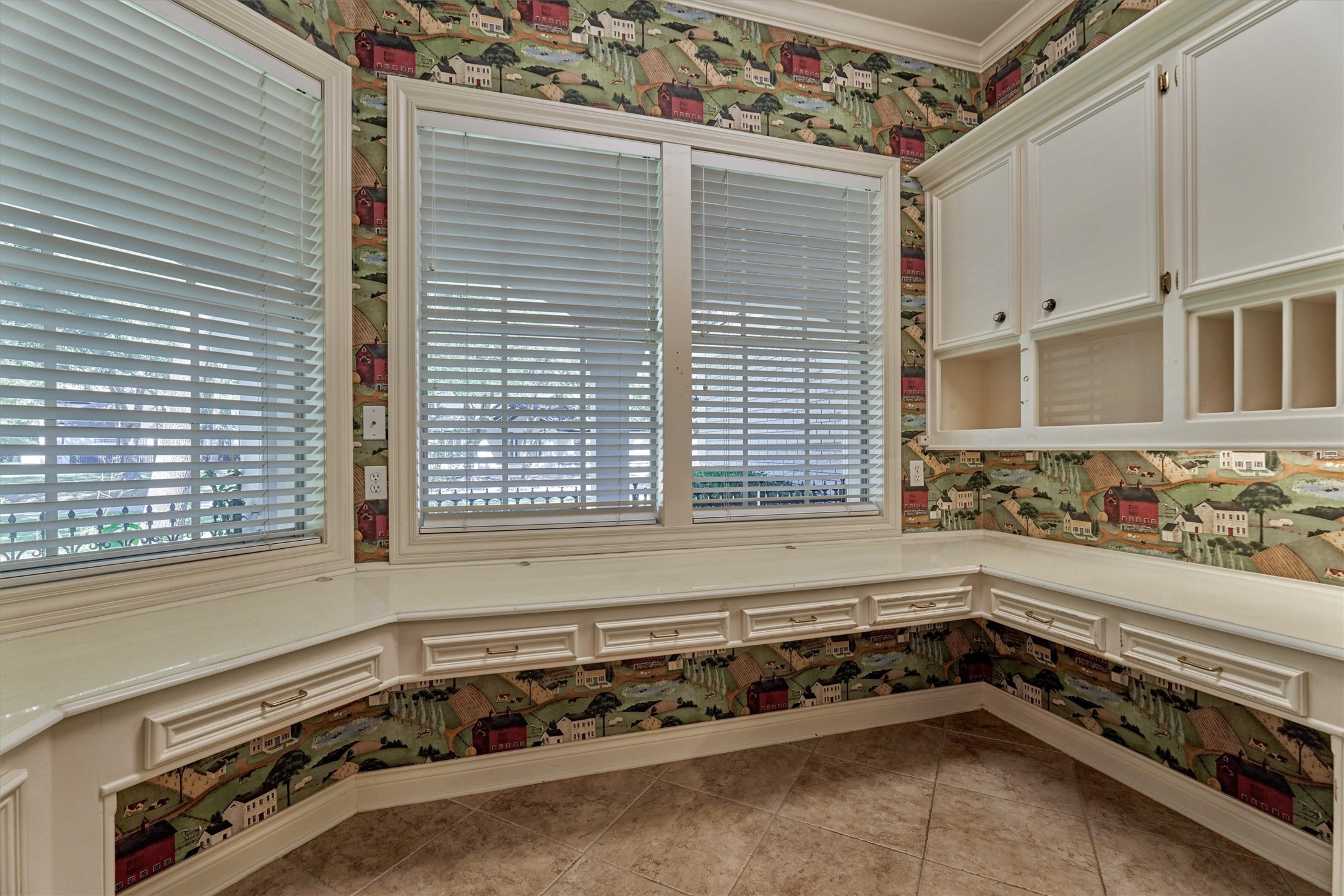 Another view of the utility room shows a built-in L-shaped counter that works as a desk, craft table, or folding station. A row of raised-panel drawers with brass pulls runs beneath the counter, and upper cabinets with open cubbies and vertical dividers keep papers and supplies organized. A bay of windows with blinds fills the room with natural light, and crown molding and tile floors add polished detail.