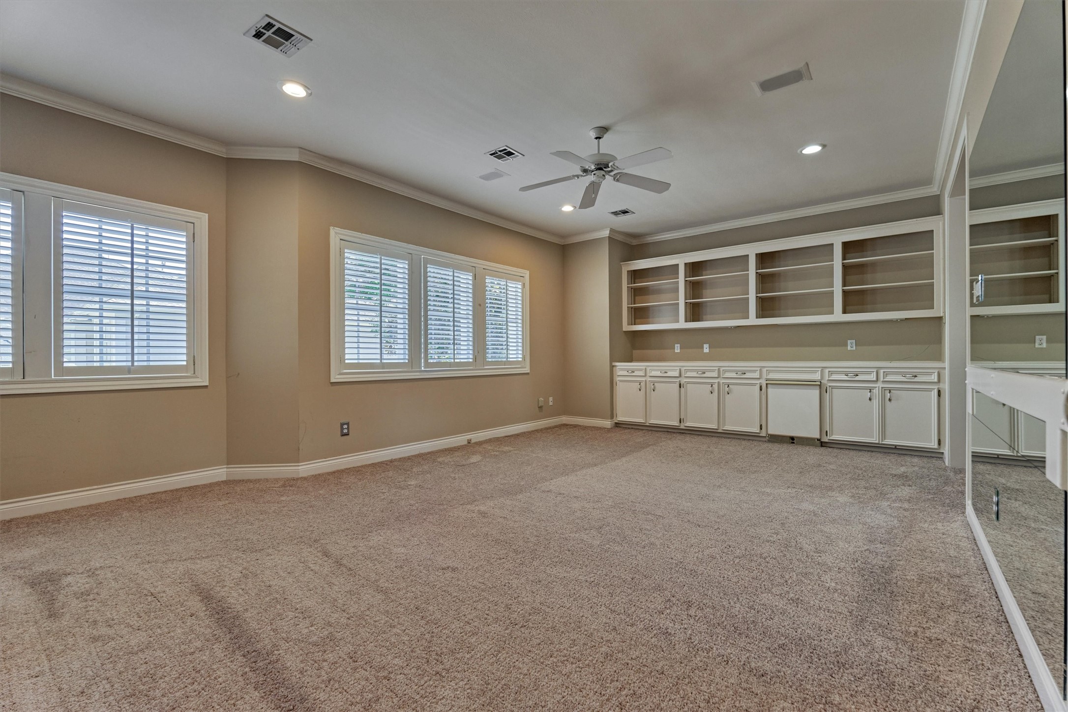 This upstairs game room is a large, open space with crown molding, recessed lighting, and a ceiling fan. A full wall of white built-ins has open shelving above and raised-panel cabinets below, with a counter that has room for a mini fridge or beverage center. A wide bank of plantation-shuttered windows lets in plenty of natural light, and a mirrored wall at the right edge adds depth. The Jack and Jill bath is steps away, and plush carpet runs throughout.