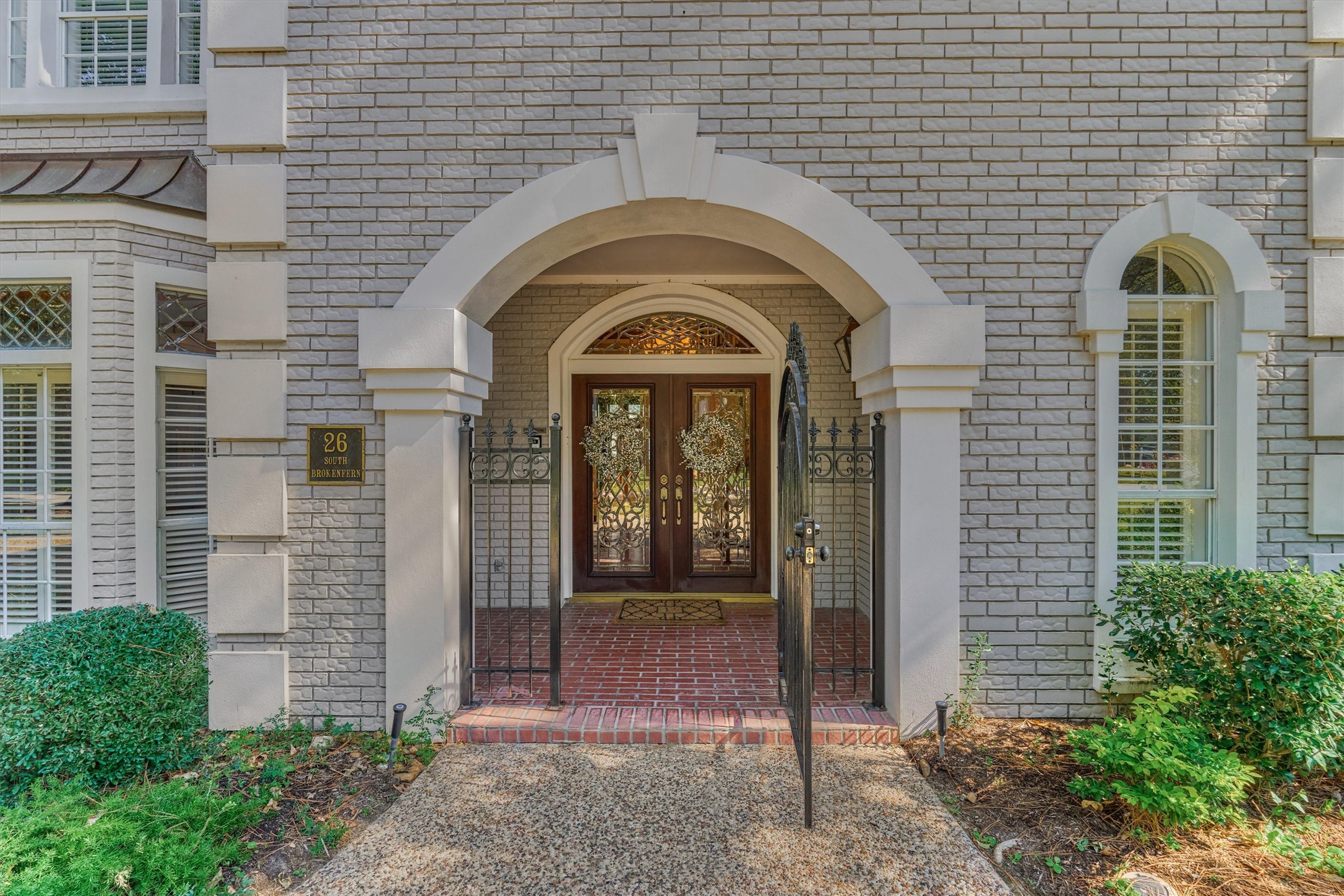 A grand stucco arch with a keystone-style accent frames the entry to this brick and stucco home. A wrought iron gate with scrollwork opens to a brick-paved porch and double mahogany-toned doors with beveled, decorative iron glass, topped by an arched transom. An arched window with stucco surround and a lantern-style light complete the elegant first impression.