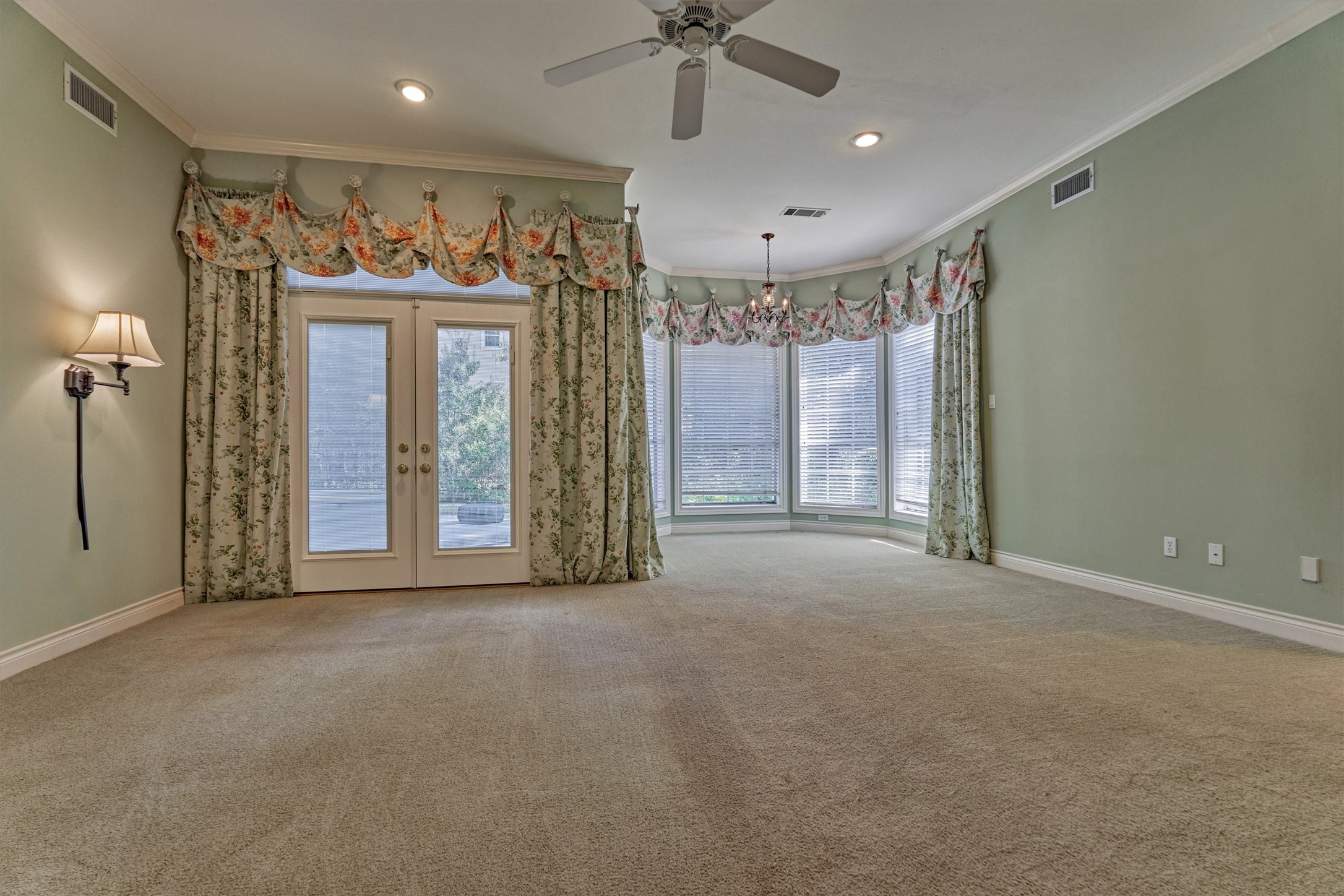 This Primary bedroom has 10-foot ceilings with deep crown molding, recessed lighting, and a ceiling fan. A bay of windows with a small chandelier makes a bright sitting or reading nook, and French doors with a transom open toward the outdoors. Plush carpet and generous proportions leave plenty of room for a king-size bed and a full seating area, and the room overlooks the backyard.