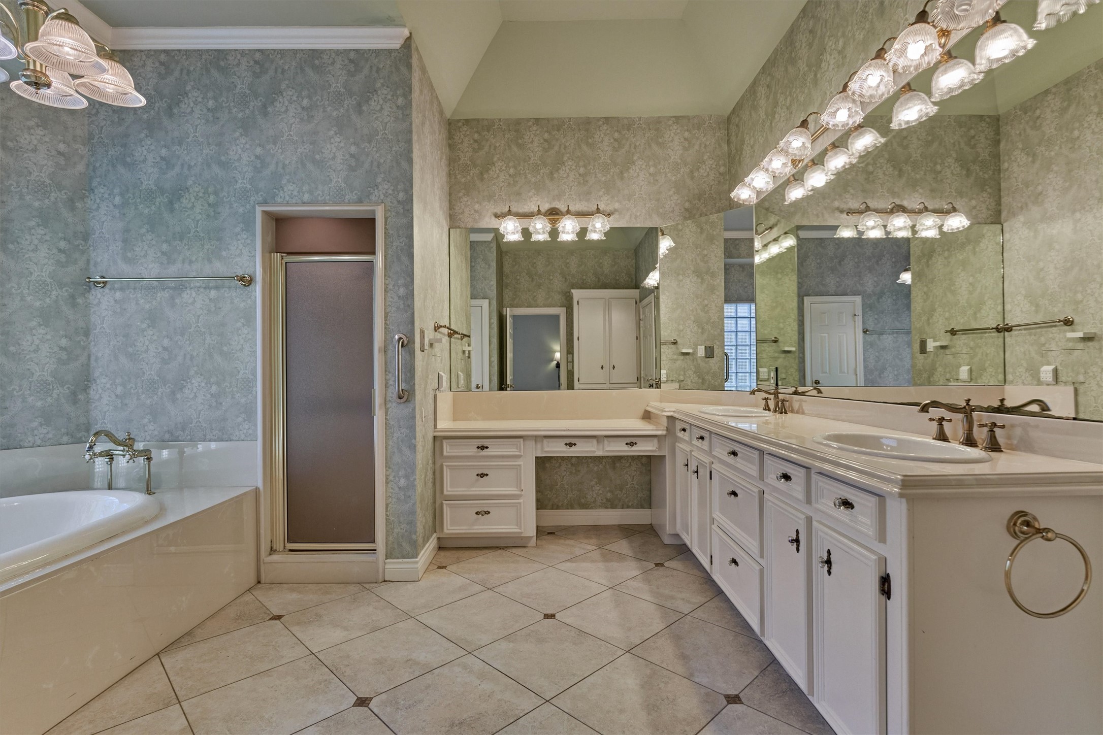 This primary bath has a vaulted ceiling and a diagonal-set tile floor with dark accent inserts. A double-sink vanity with raised-panel cabinetry, crystal-style knobs, and bronze-finish faucets sits beneath a wall of mirrors, and a separate makeup vanity with drawers adds a seated dressing area. A drop-in soaking tub with a vintage-style deck faucet and a separate walk-in shower complete the room, along with a private water closet and a glass block window.
