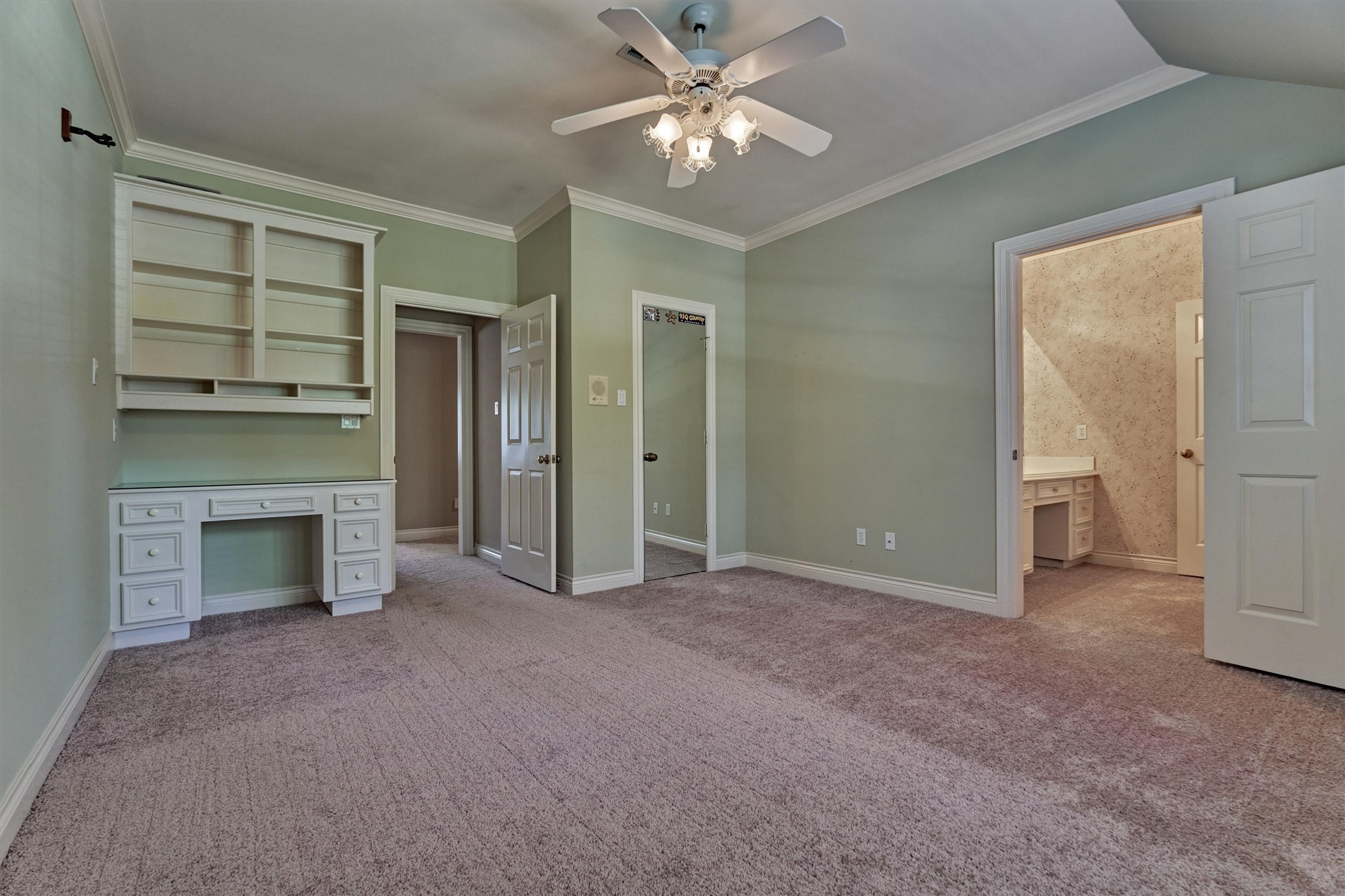 This second upstairs bedroom has a built-in desk with drawers and an upper hutch with open shelves and cubbies, perfect for homework or a computer station. Crown molding, a ceiling fan with lights, and a slightly vaulted ceiling line add detail, and plush carpet runs throughout. A closet with a mirrored door sits beside the desk area. The doorway on the right opens to this bedrooms private sink and counter area, which connects to the shared bath.