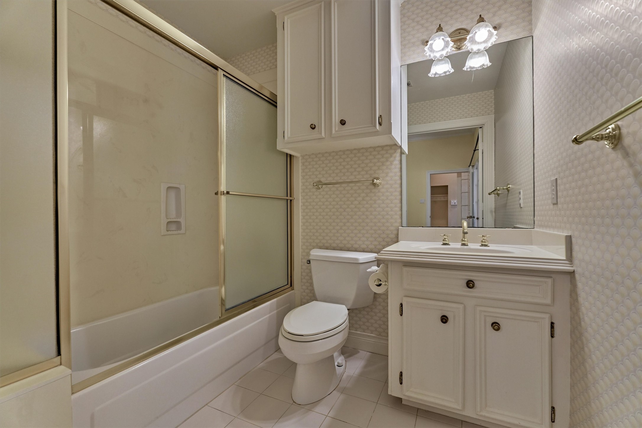 This private bath in the secondary suite is convenient and comfortable. A tub and shower combination, a single-sink vanity with cabinet storage, and extra overhead cabinets keep everything within reach. Bright, easy-care tile floors and a mirror with a multi-light fixture make it a practical space for guests, in-laws, or family.