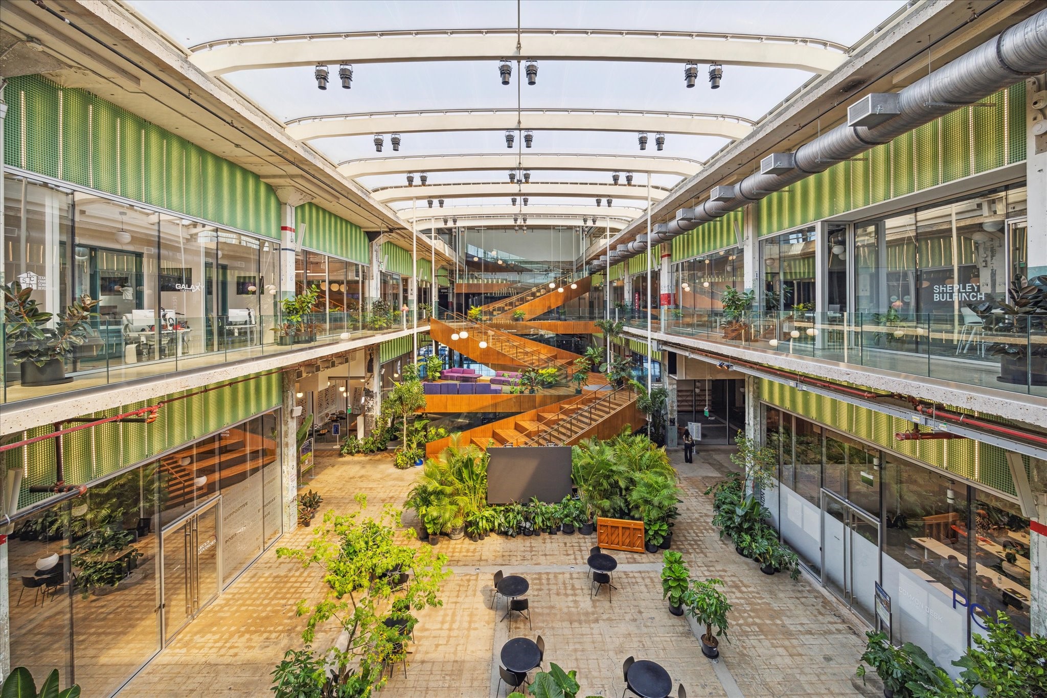 Experience vibrant modern architecture in this open-concept space, featuring a central atrium nearby with lush greenery, contemporary glass facades, and a striking wooden staircase. Perfect for those who value style and functionality.