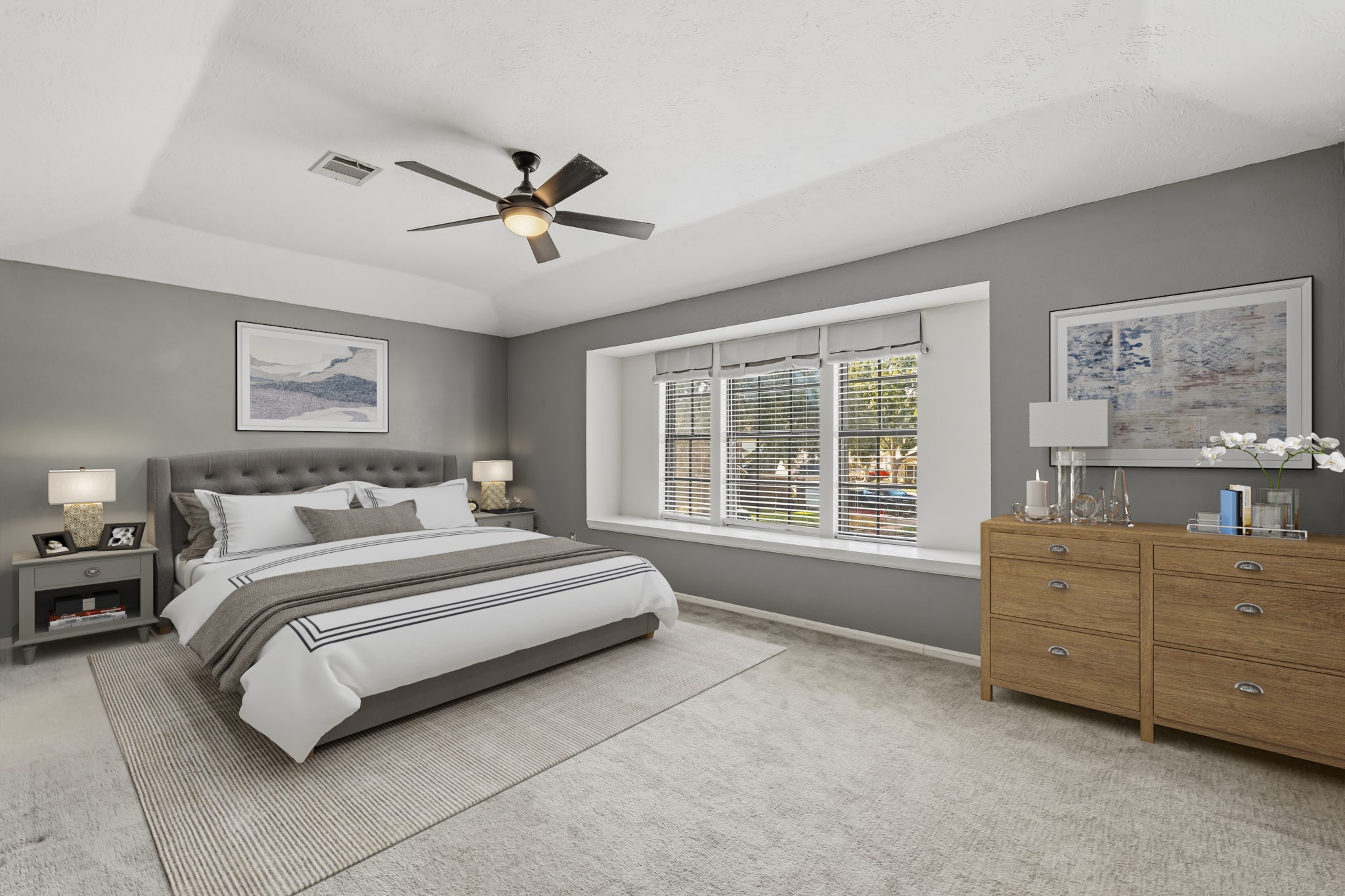 Virtually Staged: This spacious primary bedroom offers a relaxing retreat with soft neutral tones, plush carpeting, and a tray ceiling with a modern ceiling fan. A large trio of windows fills the room with natural light, while the generous floor plan provides plenty of space for a full bedroom suite and additional seating or furniture.