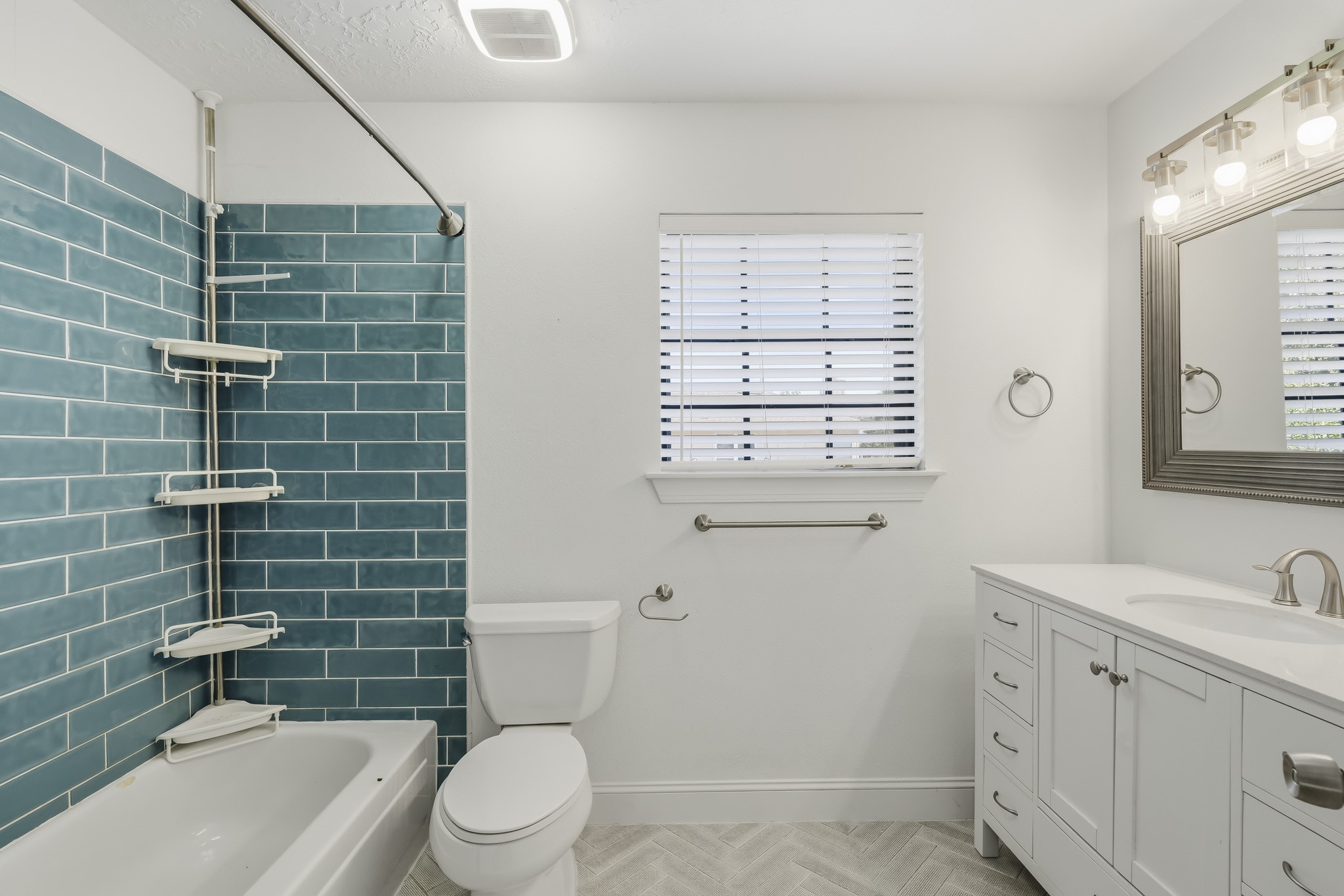 This bright full bathroom features a tub-and-shower combination accented by striking blue subway tile for a fresh pop of color. A white vanity offers generous storage and counter space, while a large framed mirror, modern lighting, and a window provide a bright, airy feel. Neutral patterned flooring completes the clean and inviting design.