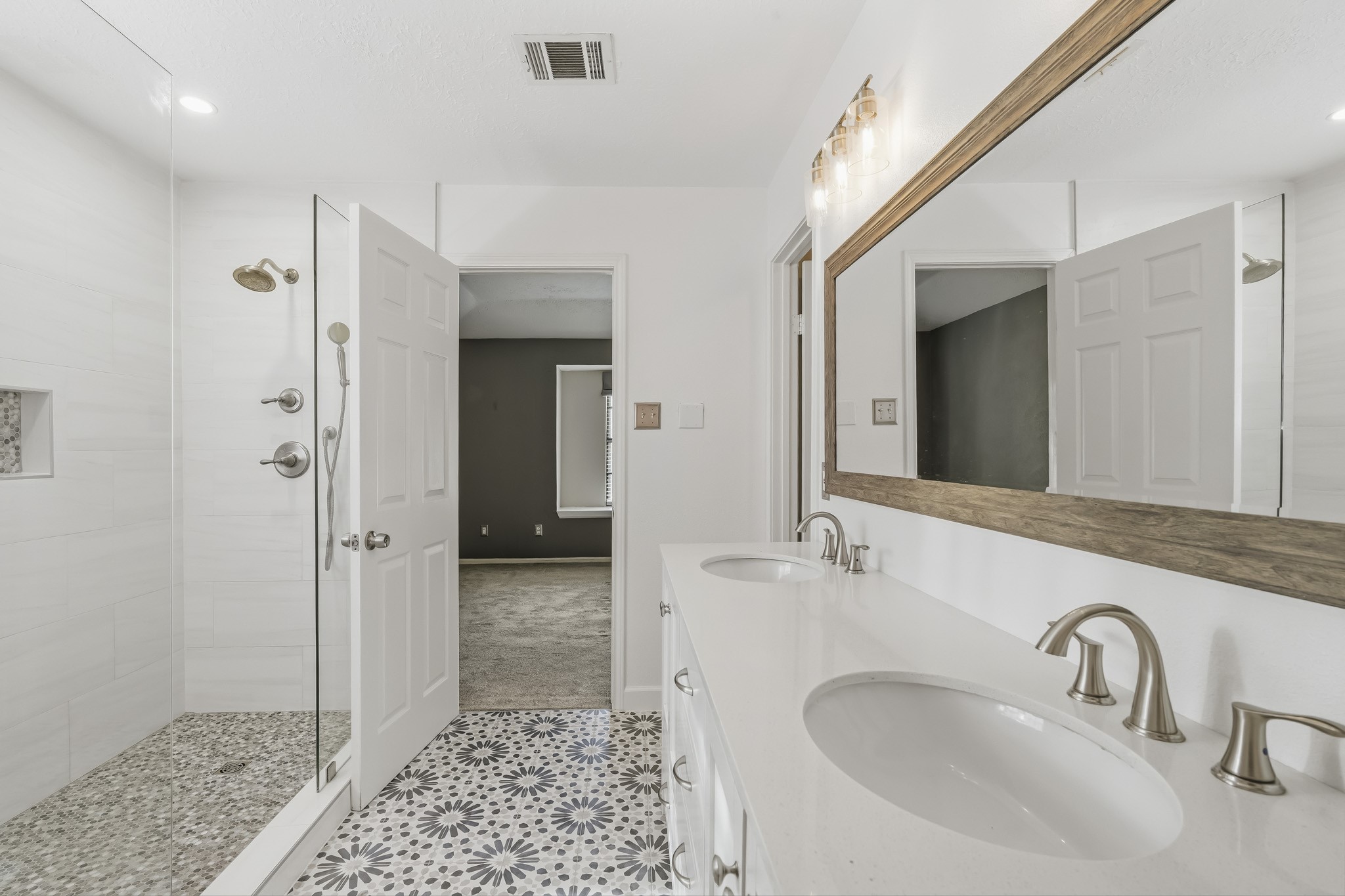 The walk-in shower offers a frameless glass enclosure, large-format tile, pebble-style flooring, and a rainfall showerhead with handheld fixture. Eye-catching patterned tile flooring adds character, while bright neutral finishes create a fresh, modern feel.