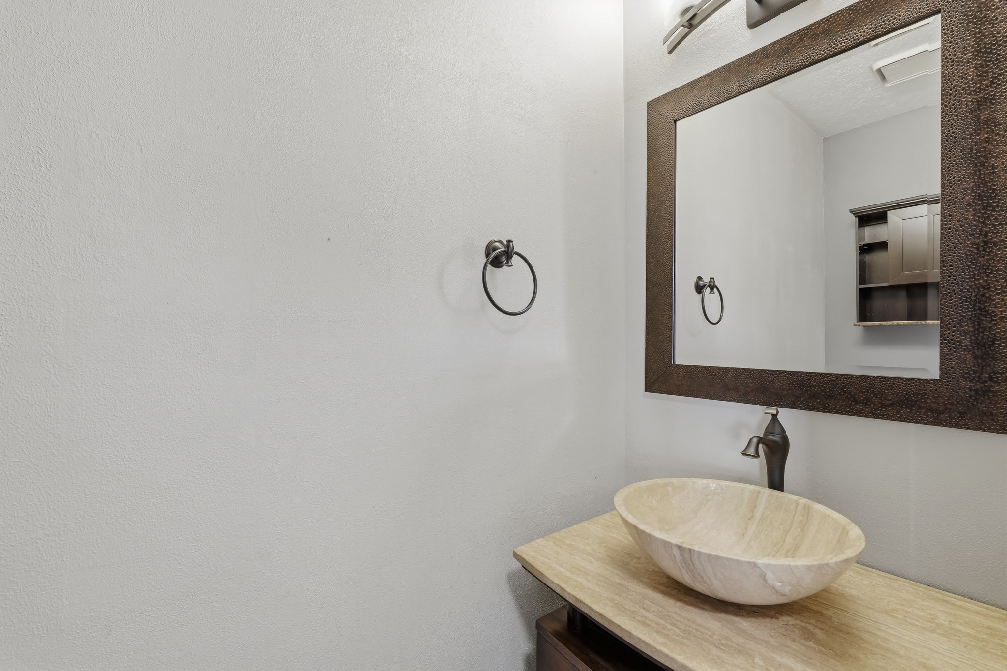 This stylish half bath features a distinctive stone vessel sink paired with a dark bronze faucet and a warm-toned countertop. An oversized framed mirror, coordinating bronze fixtures, and neutral finishes create a polished, inviting space for guests.