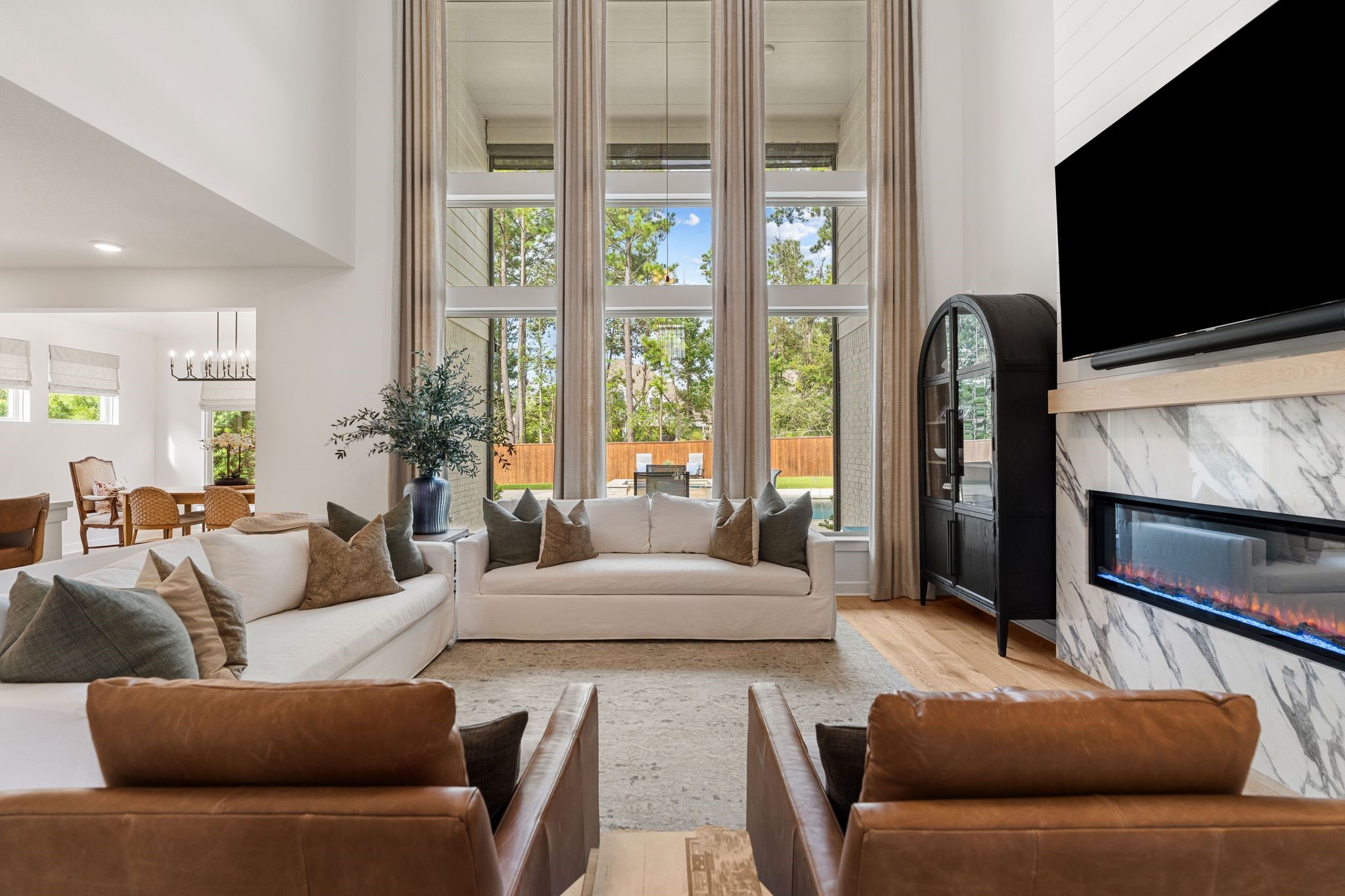 Breathtaking two-story family room showcasing soaring ceilings, dramatic floor-to-ceiling windows with custom drapery, and a stunning contemporary fireplace with elegant marble surround. Beautiful natural light and picturesque views of the private backyard oasis create a luxurious yet inviting space perfect for entertaining and everyday living!