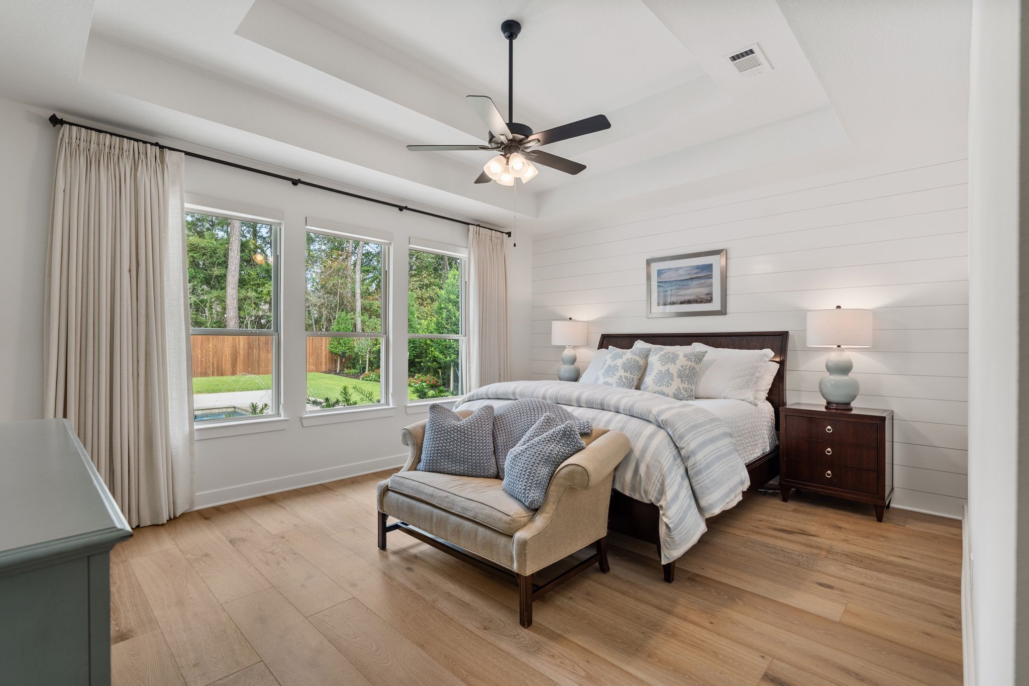 Serene primary suite featuring elegant tray ceilings, beautiful shiplap accent wall, warm wood flooring, and expansive windows overlooking the private backyard oasis and lush greenbelt.
