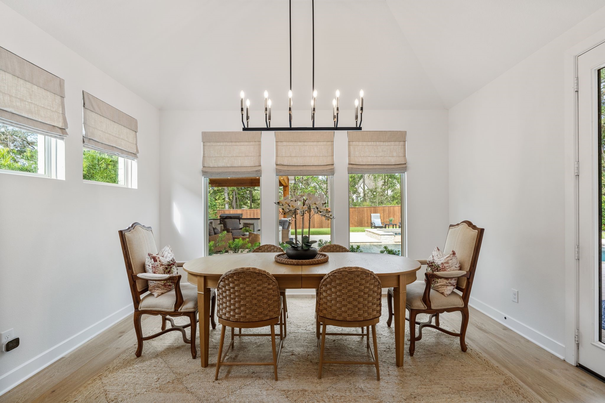 Dining area featuring a stunning designer chandelier, 9" white oak flooring, custom Roman shades, and walls of windows showcasing picturesque views of the private backyard oasis.