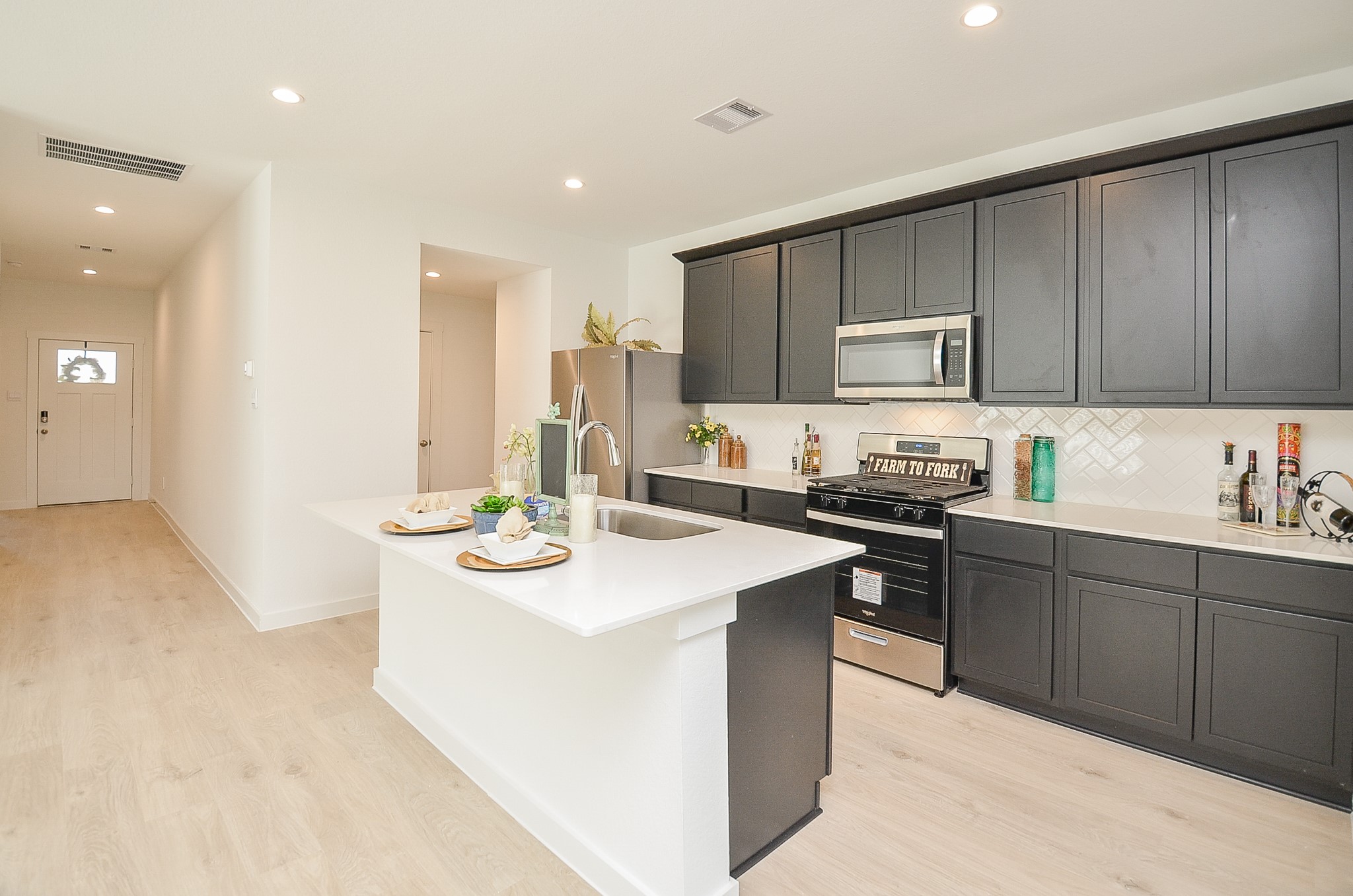 A modern kitchen featuring sleek dark cabinetry, stainless steel appliances, and a spacious island with bar seating, seamlessly connected to a bright hallway.