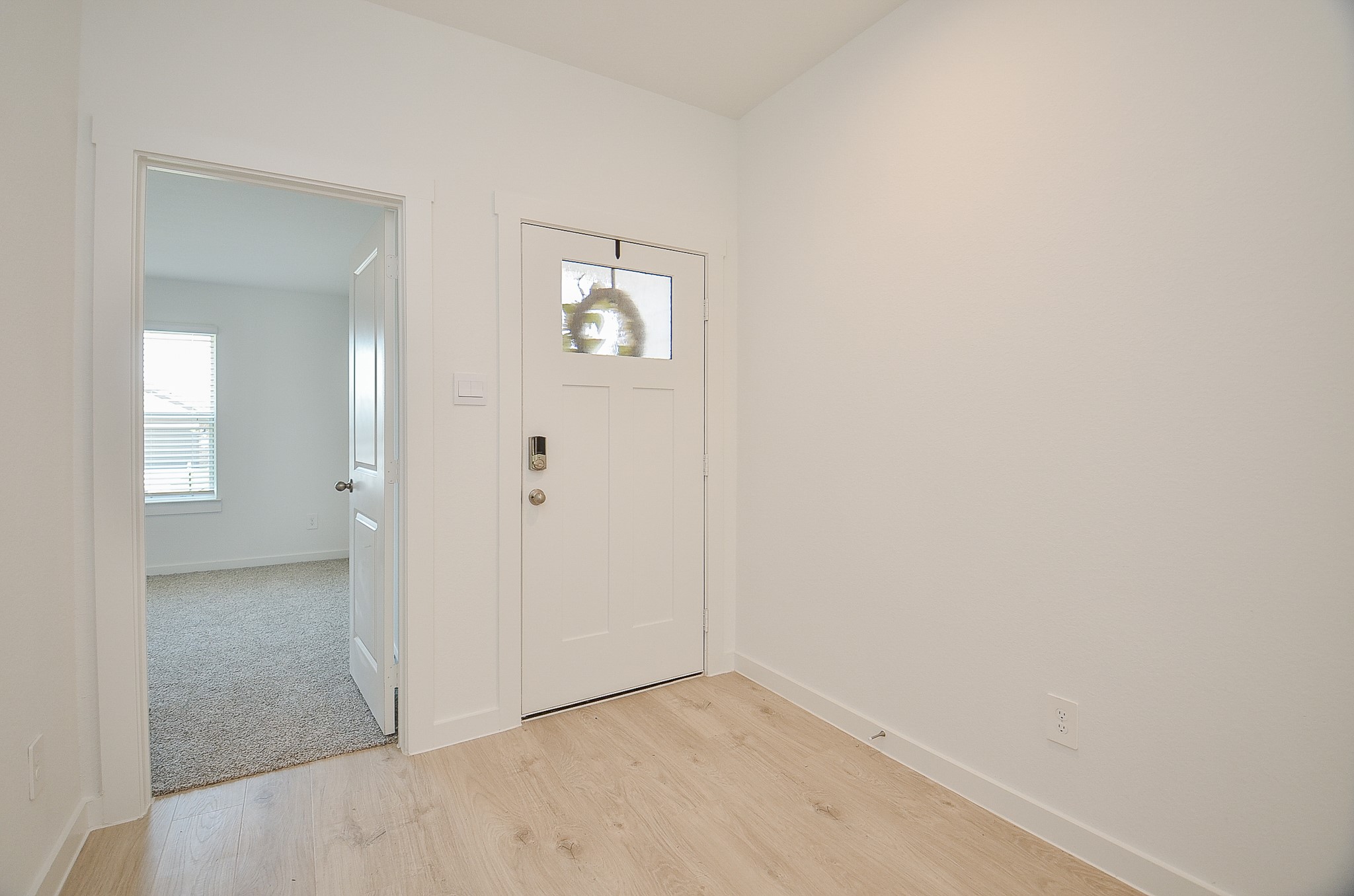 Bright and inviting entryway with light wood flooring and a classic white front door, leading into a cozy, carpeted room with ample natural light.