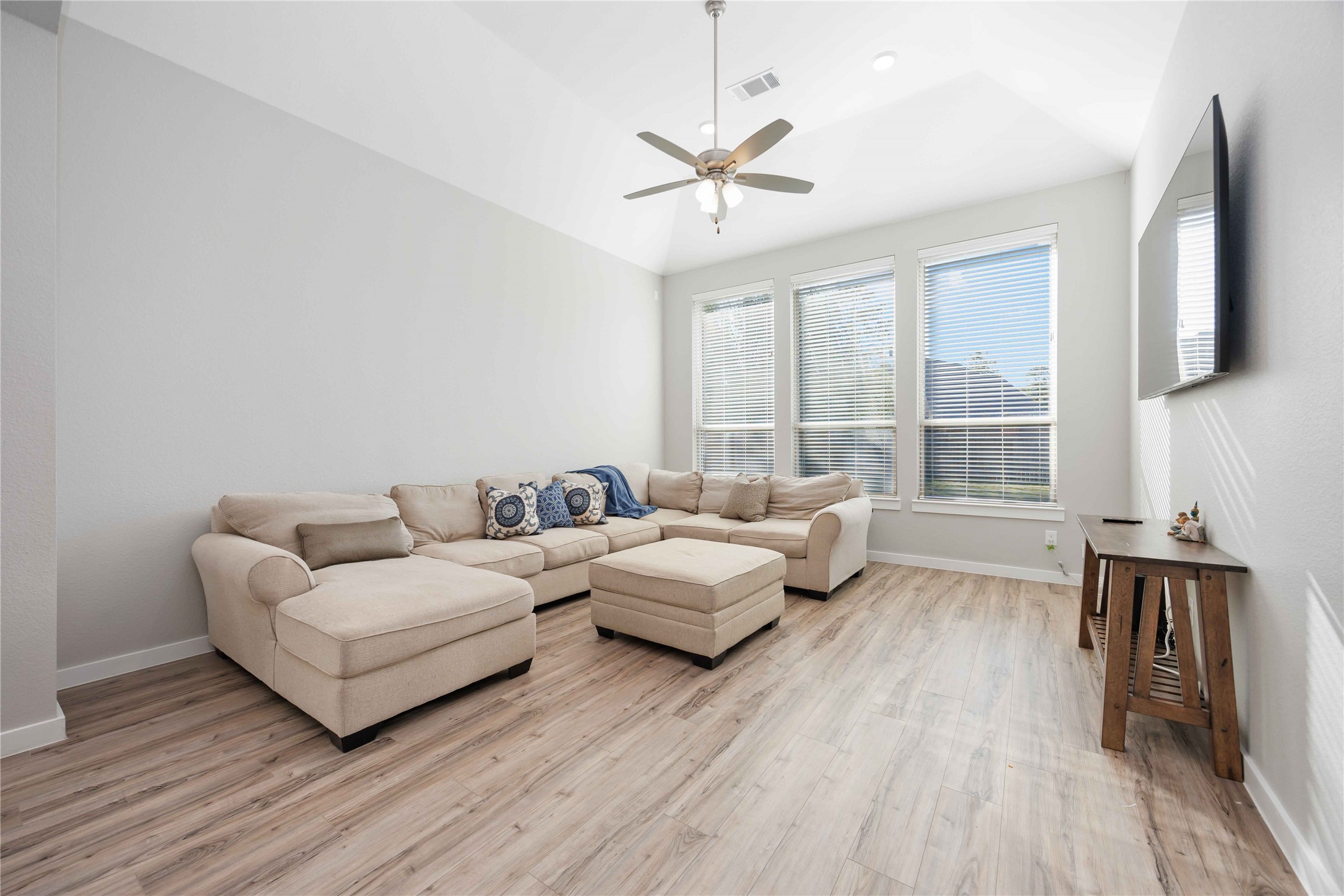 Bright and spacious living room with large windows, wood flooring, and a neutral-toned sectional sofa. Features high ceilings, a ceiling fan, and a wall-mounted TV for a modern touch.