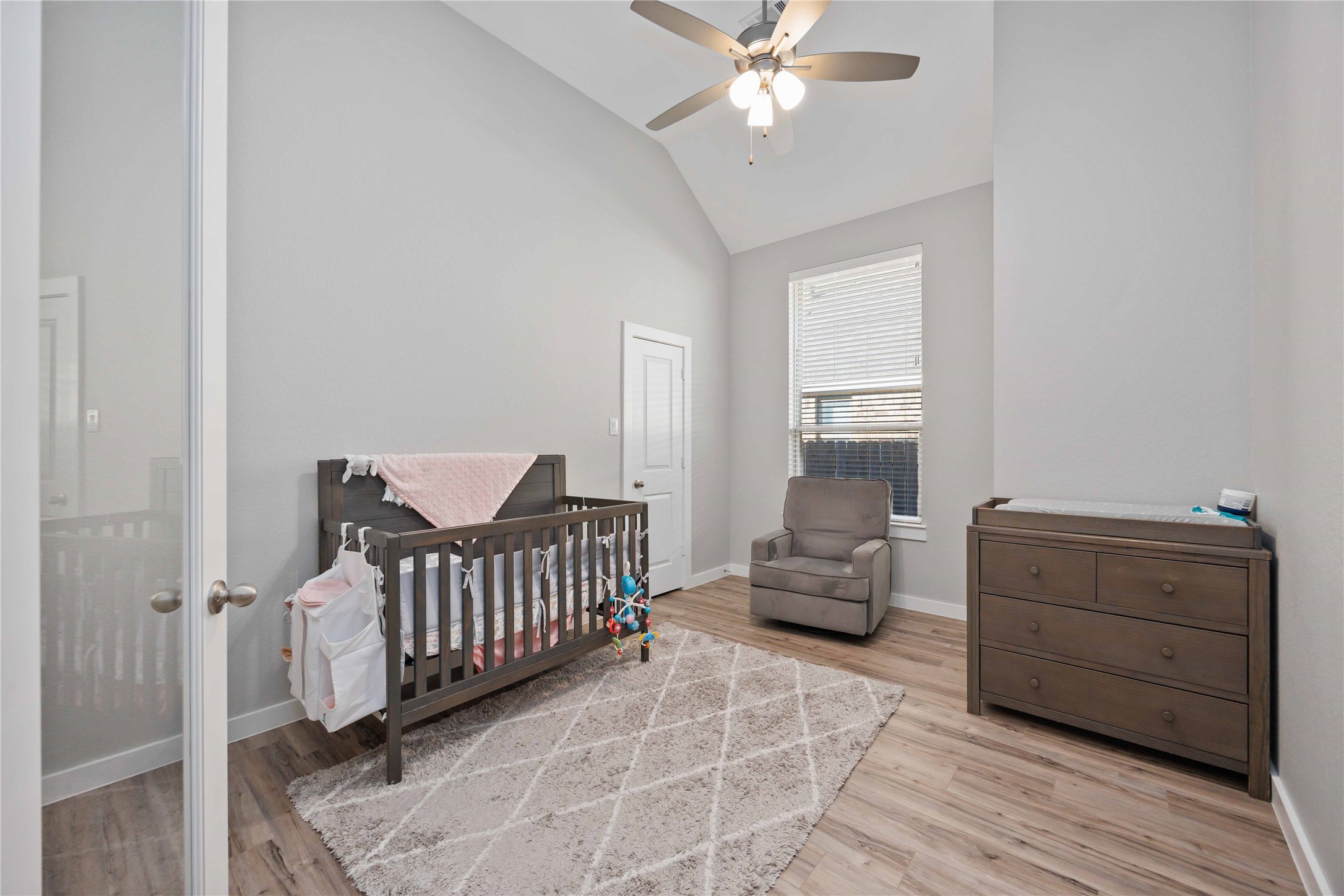 This cozy nursery features a crib, a comfortable armchair, and a dresser, all set against soft gray walls and wood flooring. A ceiling fan and large window provide ample light and ventilation.
