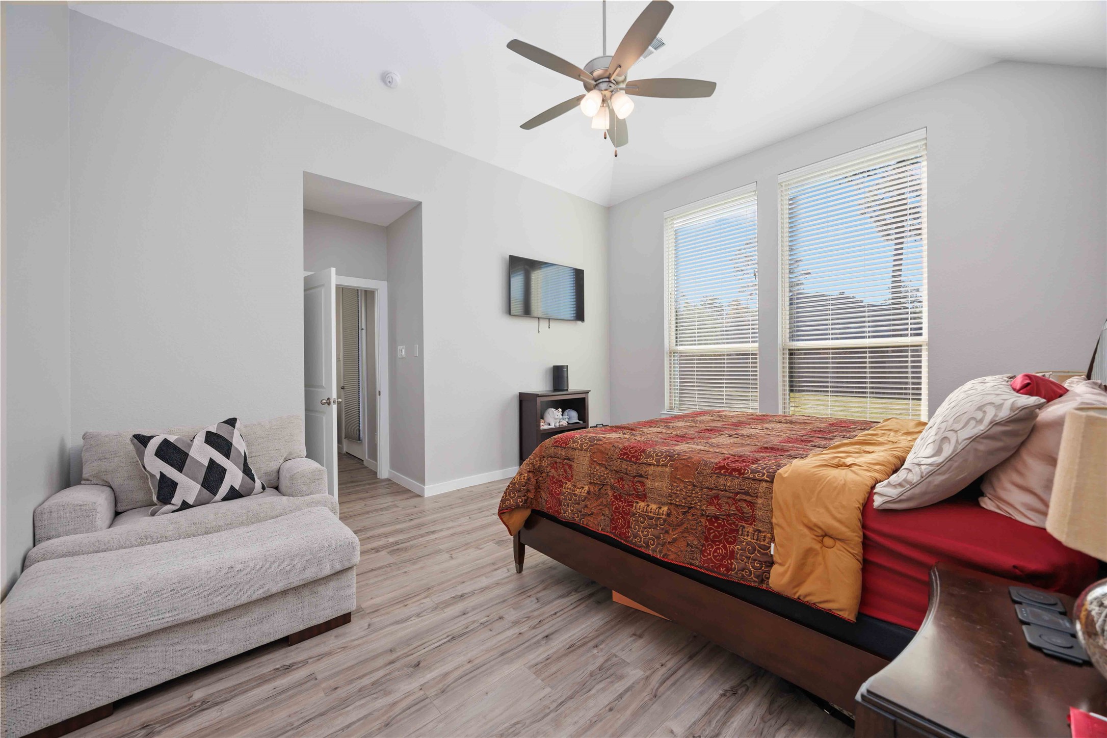 This bedroom is bright and spacious, featuring large windows, a ceiling fan, and light-colored flooring. It includes a cozy sitting area, mounted TV, and a comfortable bed, creating a welcoming and functional space.