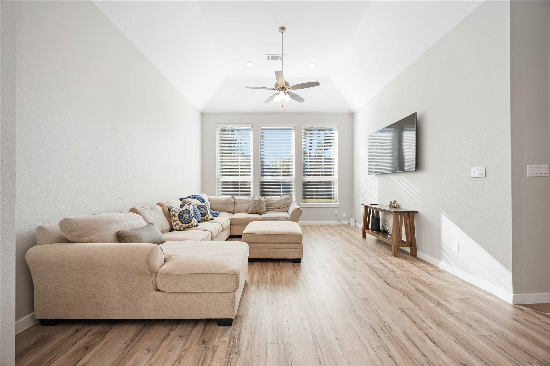 Bright and spacious living room with high ceilings, featuring a cozy beige sectional sofa, wood flooring, and a wall-mounted TV. Large windows let in plenty of natural light.