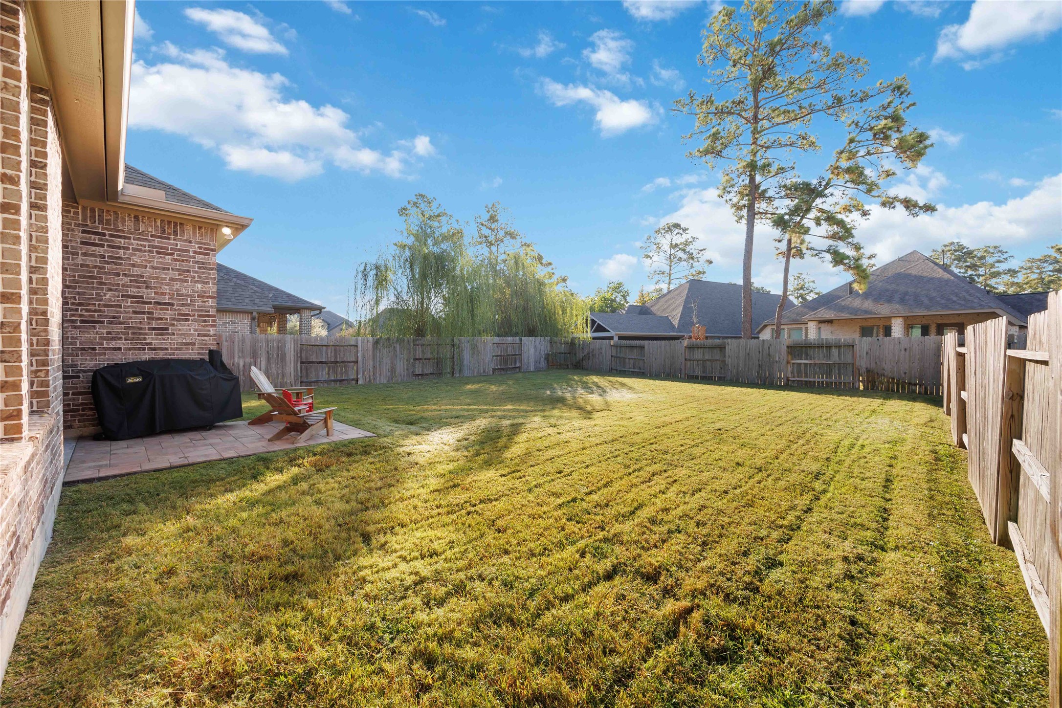 This photo shows a spacious backyard with a well-maintained lawn, bordered by a wooden fence. There's a small patio area with outdoor seating and a grill, perfect for relaxation or entertaining. The yard is surrounded by other houses, offering privacy and a serene atmosphere.
