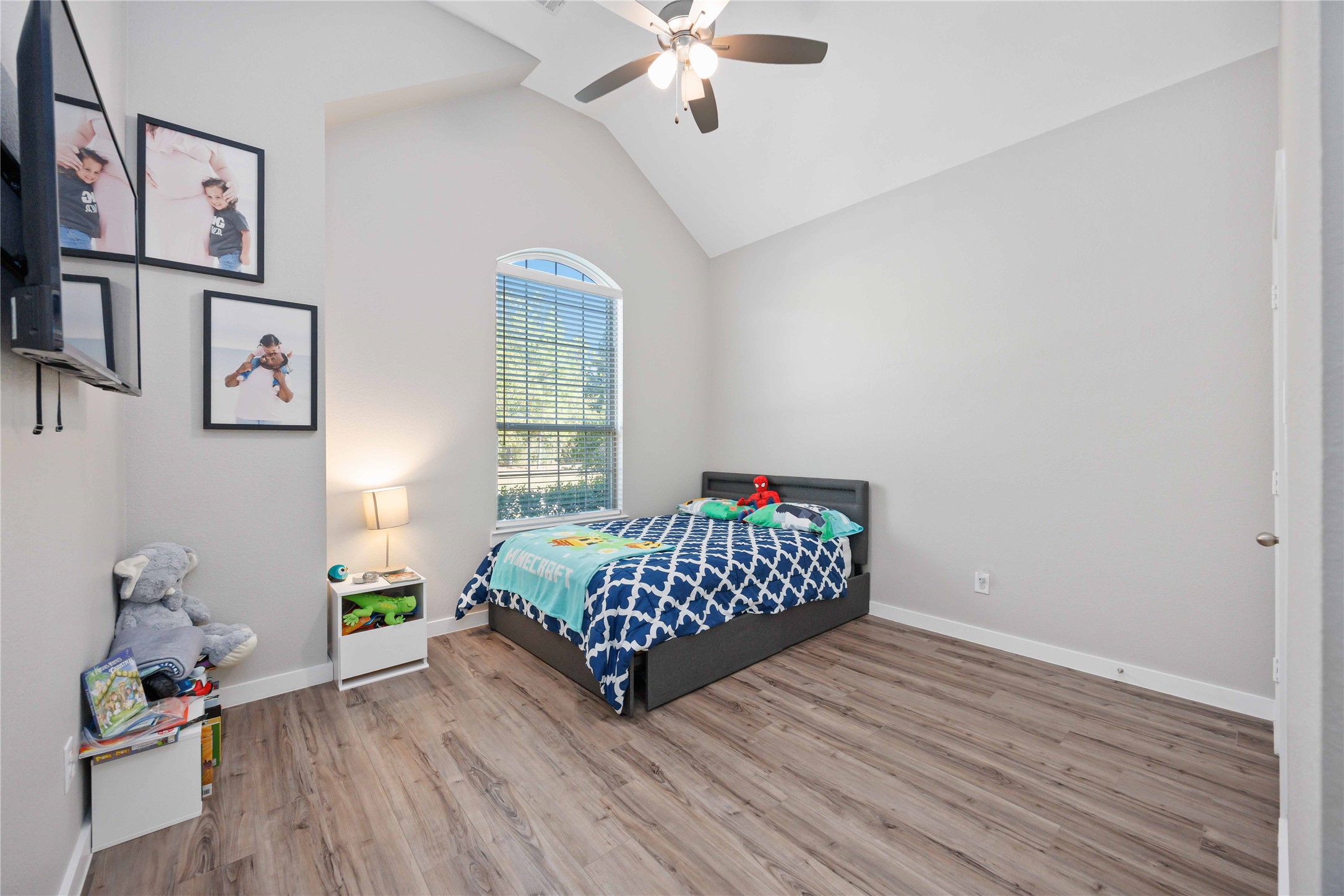 This bedroom features high ceilings and abundant natural light from a large, arched window. The room has wood-style flooring, a ceiling fan, and neutral walls, offering a modern and spacious feel. Its ideal for a child's room with playful decor and ample space for a bed and furniture.