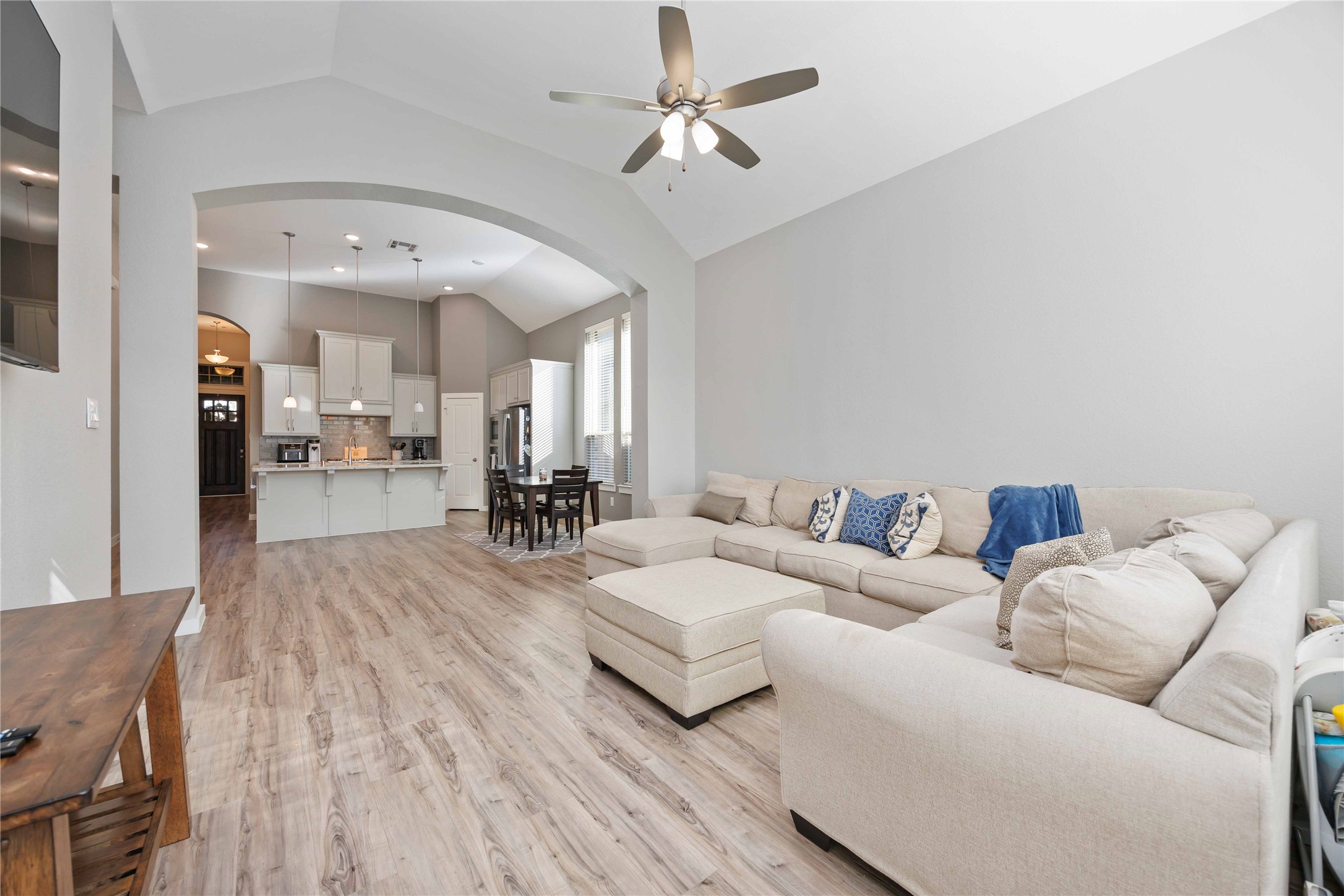 This inviting open-concept living space features a cozy beige sectional, vaulted ceilings, and wood-look flooring. It seamlessly connects to a modern kitchen with a breakfast bar and a dining area, offering a spacious and airy feel. Perfect for family gatherings and entertaining.